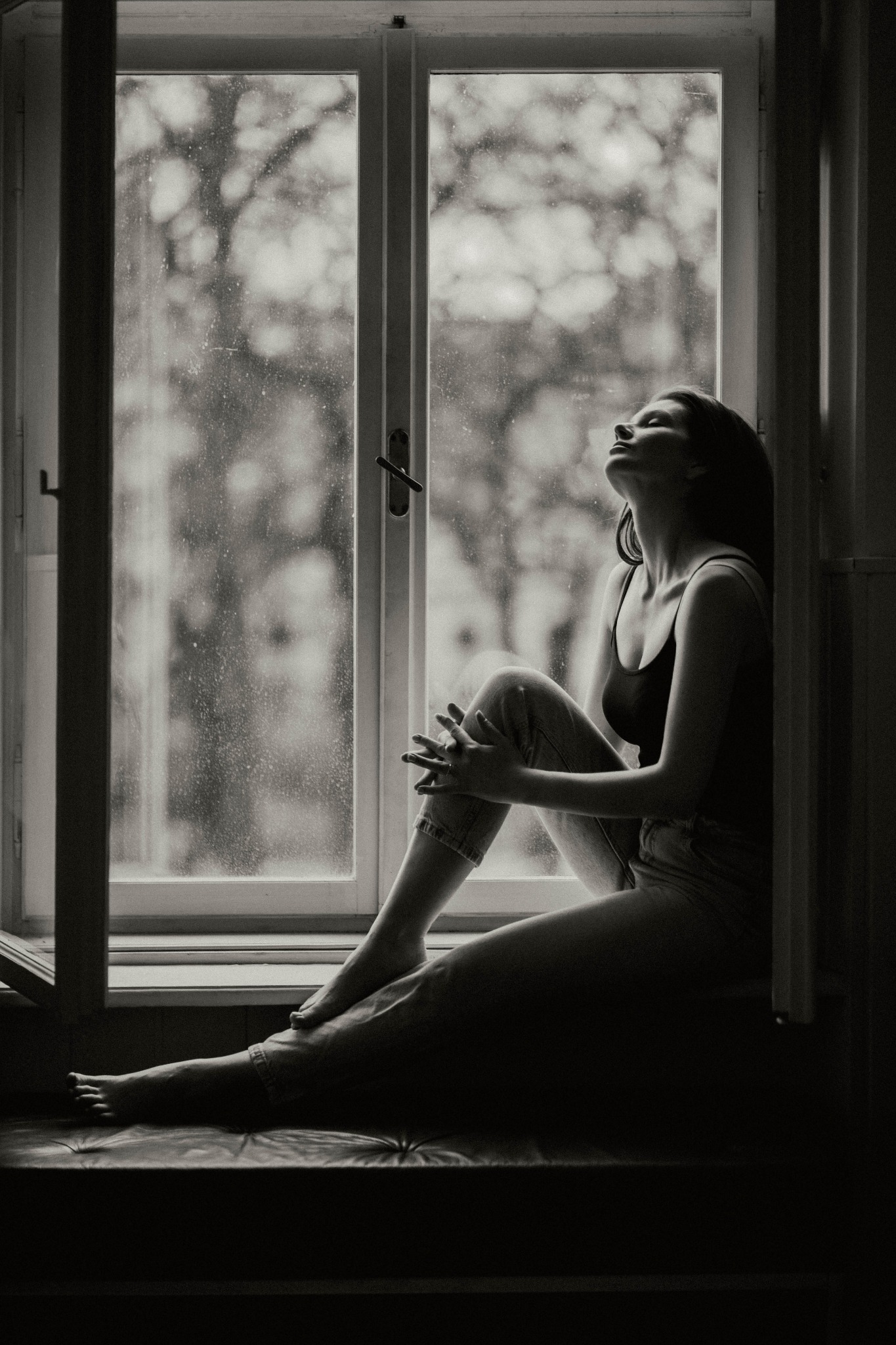 Kobieta siedząca na parapecie okna w deszczowy dzień, głowa zadarta do góry, czarno-biała fotografia artystyczna.