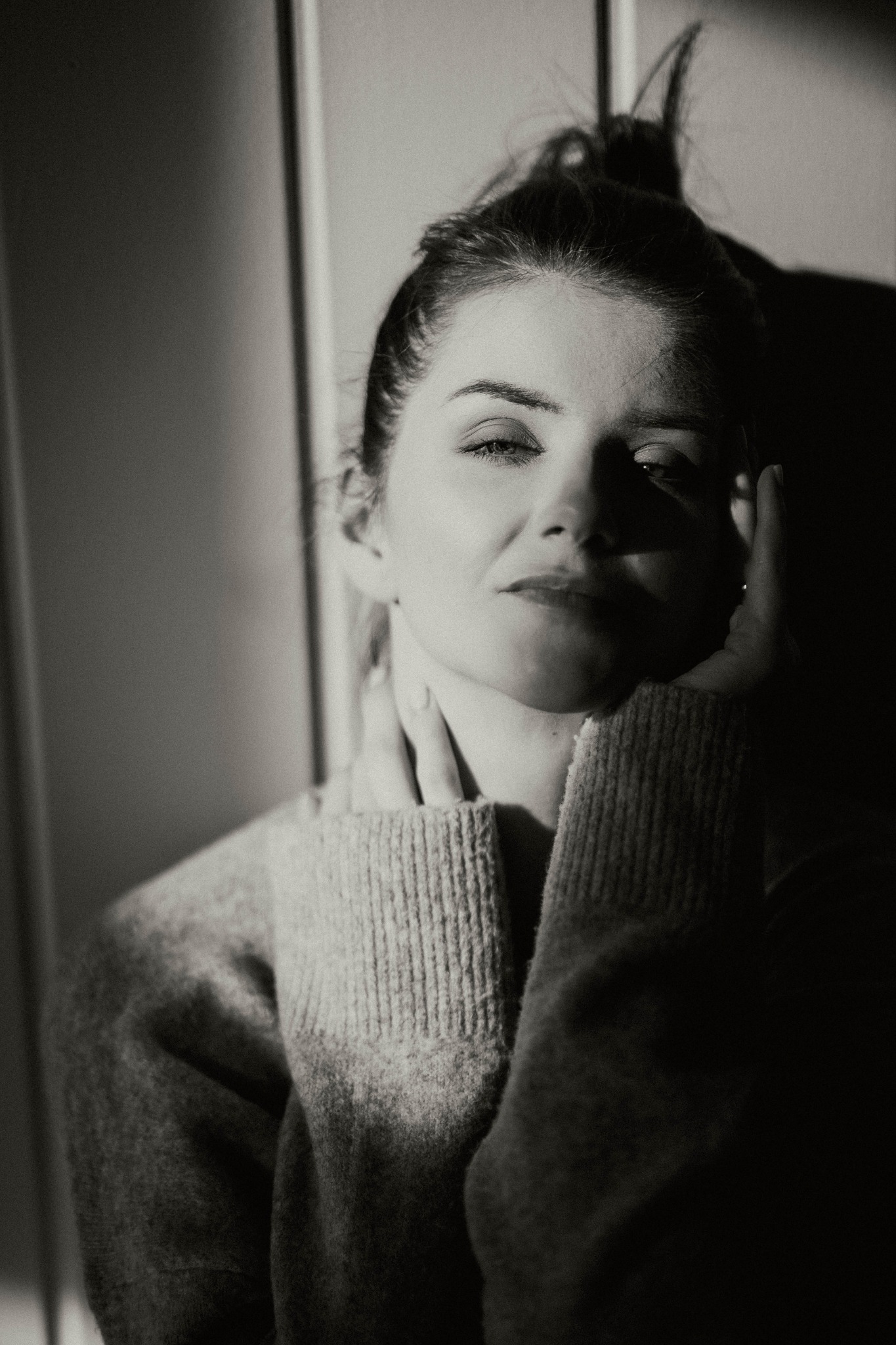 Portret kobiety w swetrze, oświetlona bocznym światłem, z rękami przy twarzy, w tonacji czarno-białej.