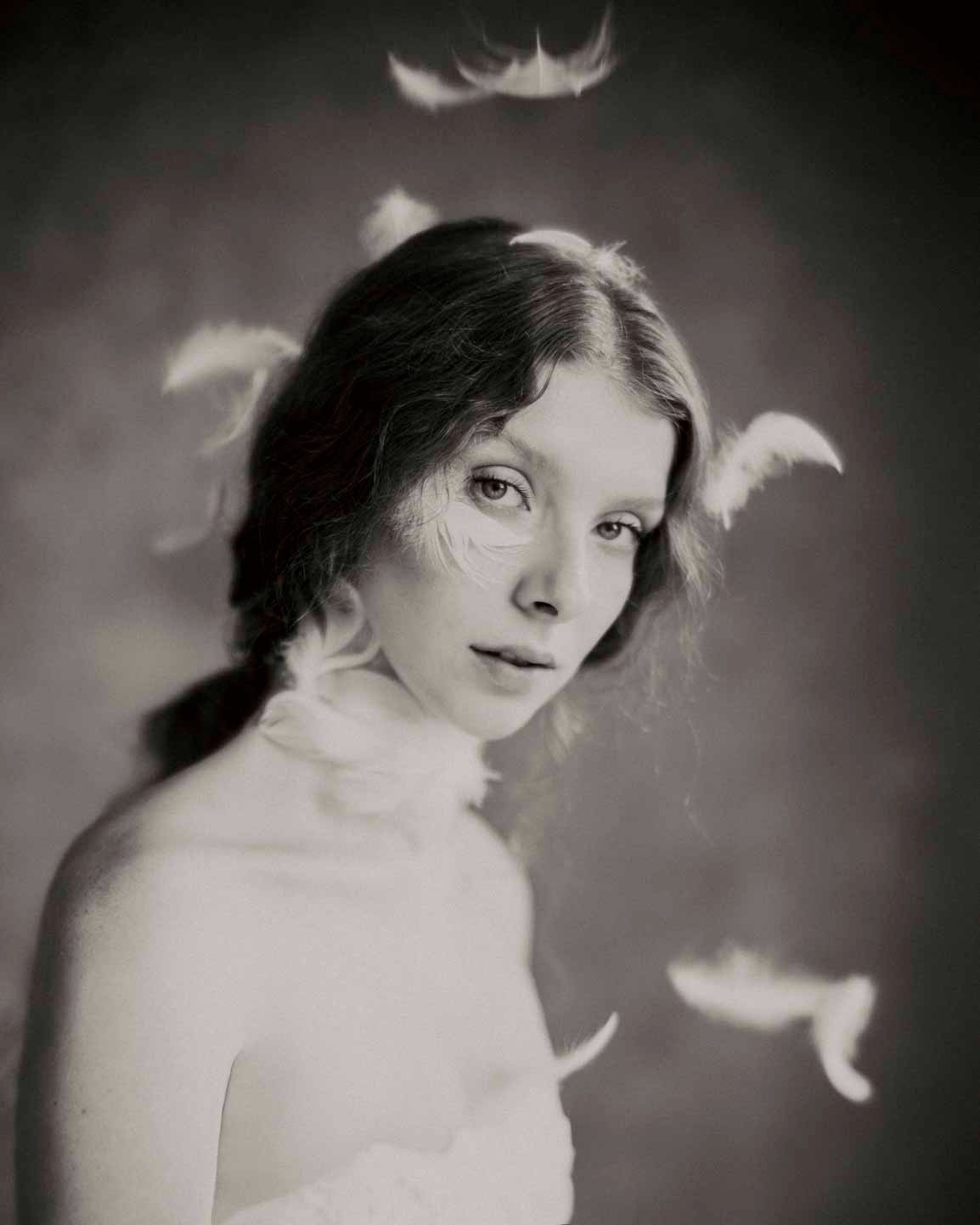 Portret kobiety z piórami wokół twarzy i na ramionach, w stylu vintage, czarno-biały, z subtelnym oświetleniem.