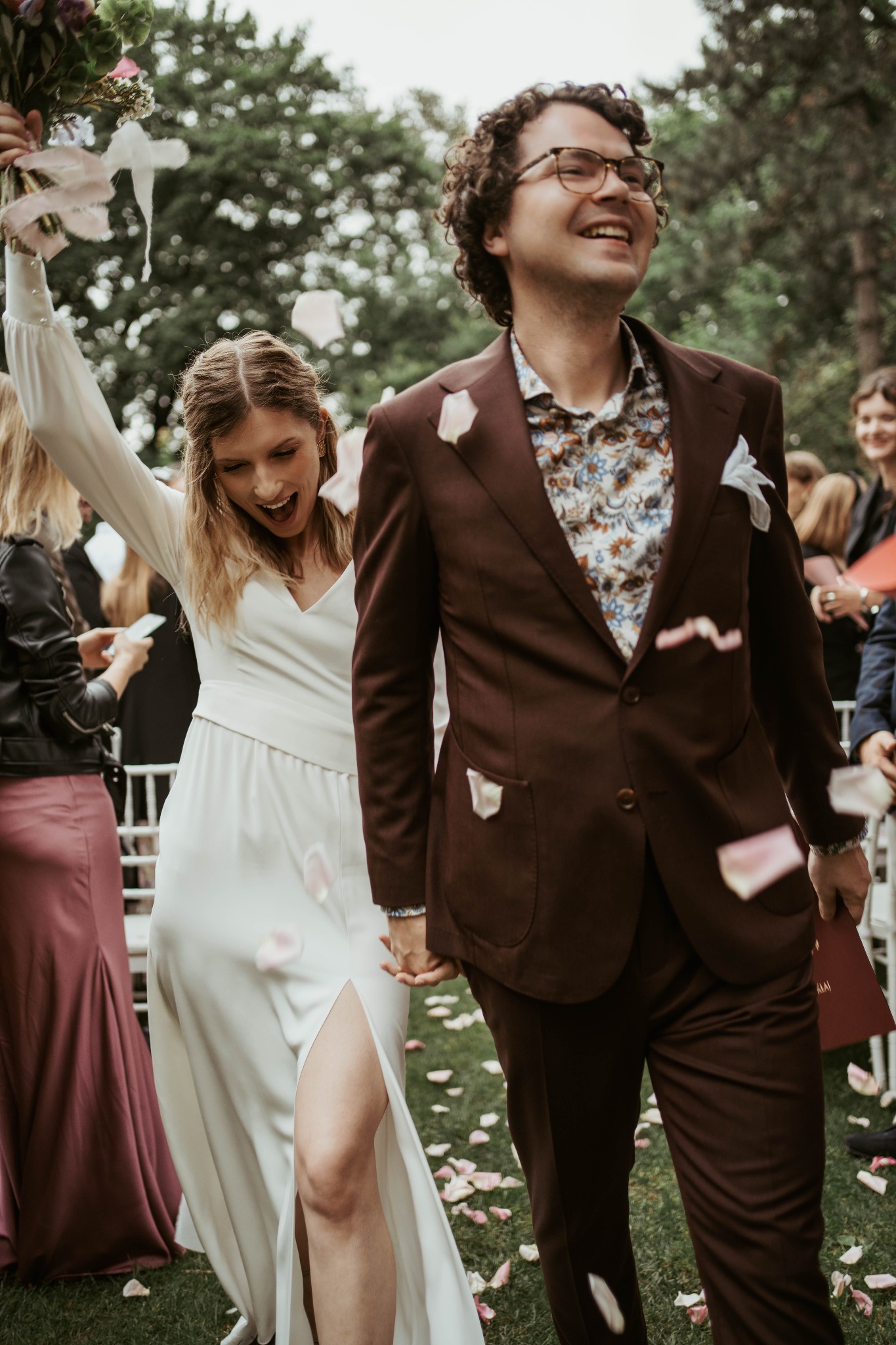 Uśmiechnięta para młoda, on w brązowym garniturze i wzorzystej koszuli, ona w białej sukni z rozcięciem, idąca przez szpaler gości obsypywana płatkami róż.