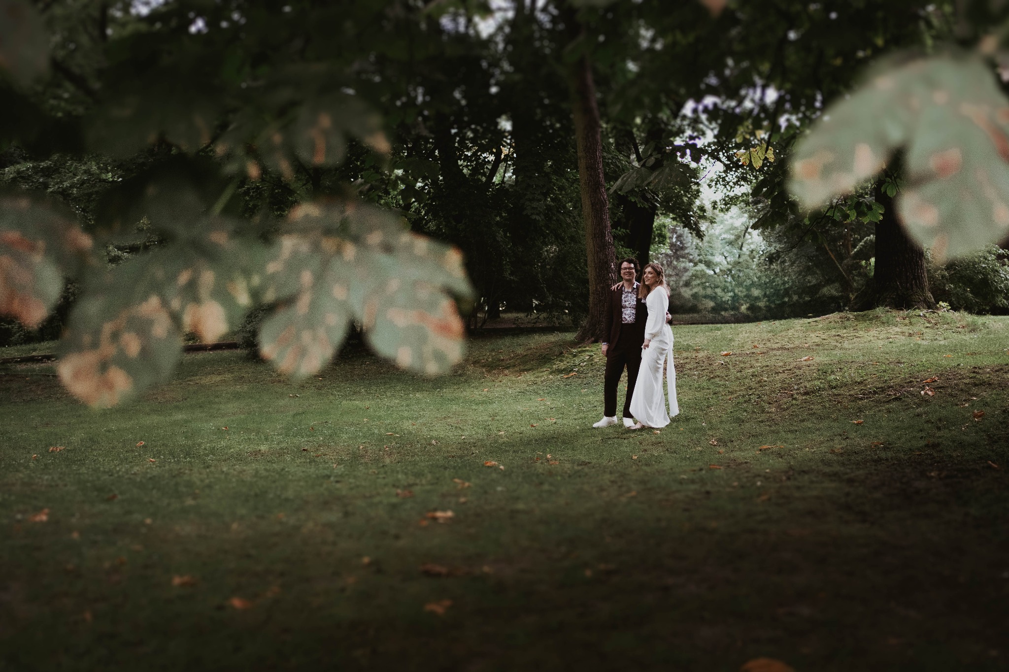 Para młoda w parku w Krakowie, panna młoda w białym kombinezonie, pan młody w brązowym garniturze w kwiaty, zdjęcie zza liści drzewa.