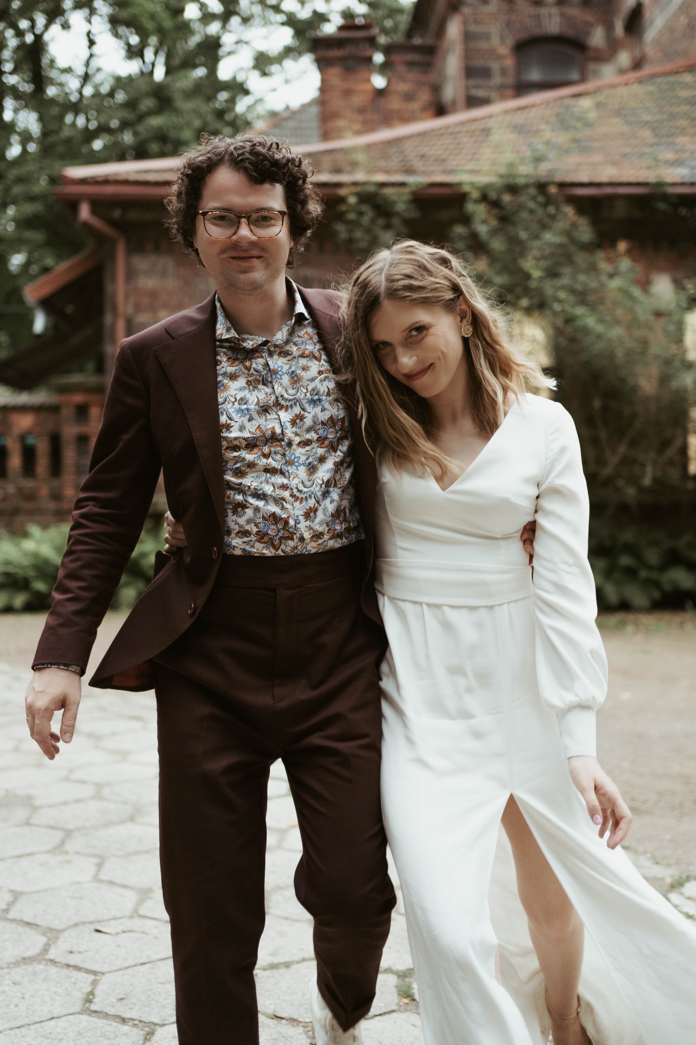 Para w eleganckich strojach spaceruje po kamiennej ścieżce na tle ceglanego budynku z zielenią. Mężczyzna w brązowym garniturze i wzorzystej koszuli, kobieta w białej sukni z rozcięciem.