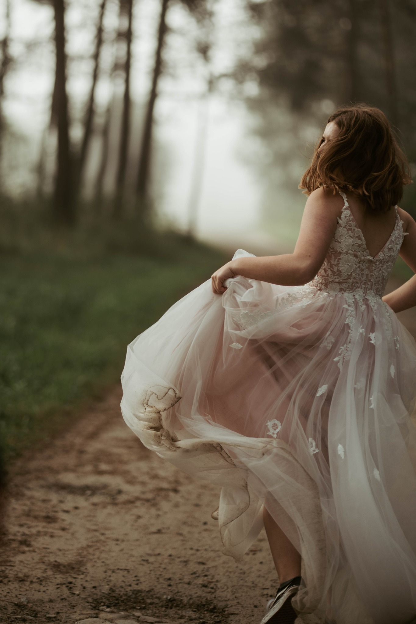 Kobieta w koronkowej sukni ślubnej z pudrowo-różowym tiulem, biegnąca leśną ścieżką w tenisówkach, unosząc brudny dół sukni.