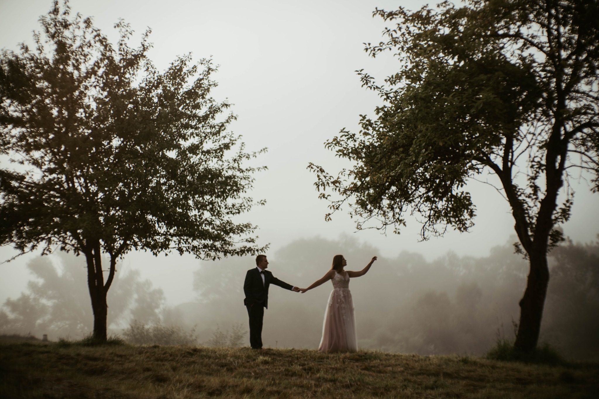 Para młoda trzymająca się za ręce, pozująca na wzgórzu w mglisty dzień, otoczona drzewami.