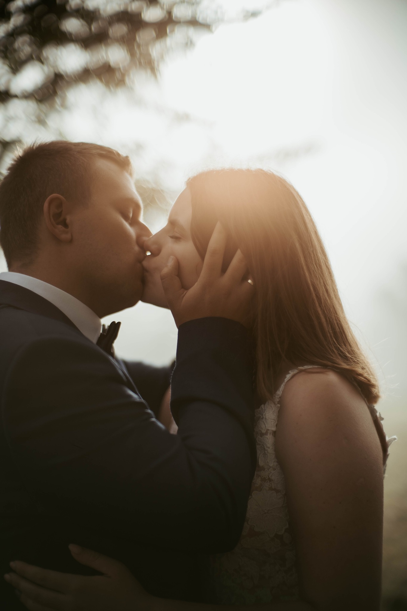 Młoda para w pocałunku podczas sesji plenerowej, on w granatowym garniturze z muchą, ona w koronkowej sukni ślubnej, delikatne światło słoneczne w tle.