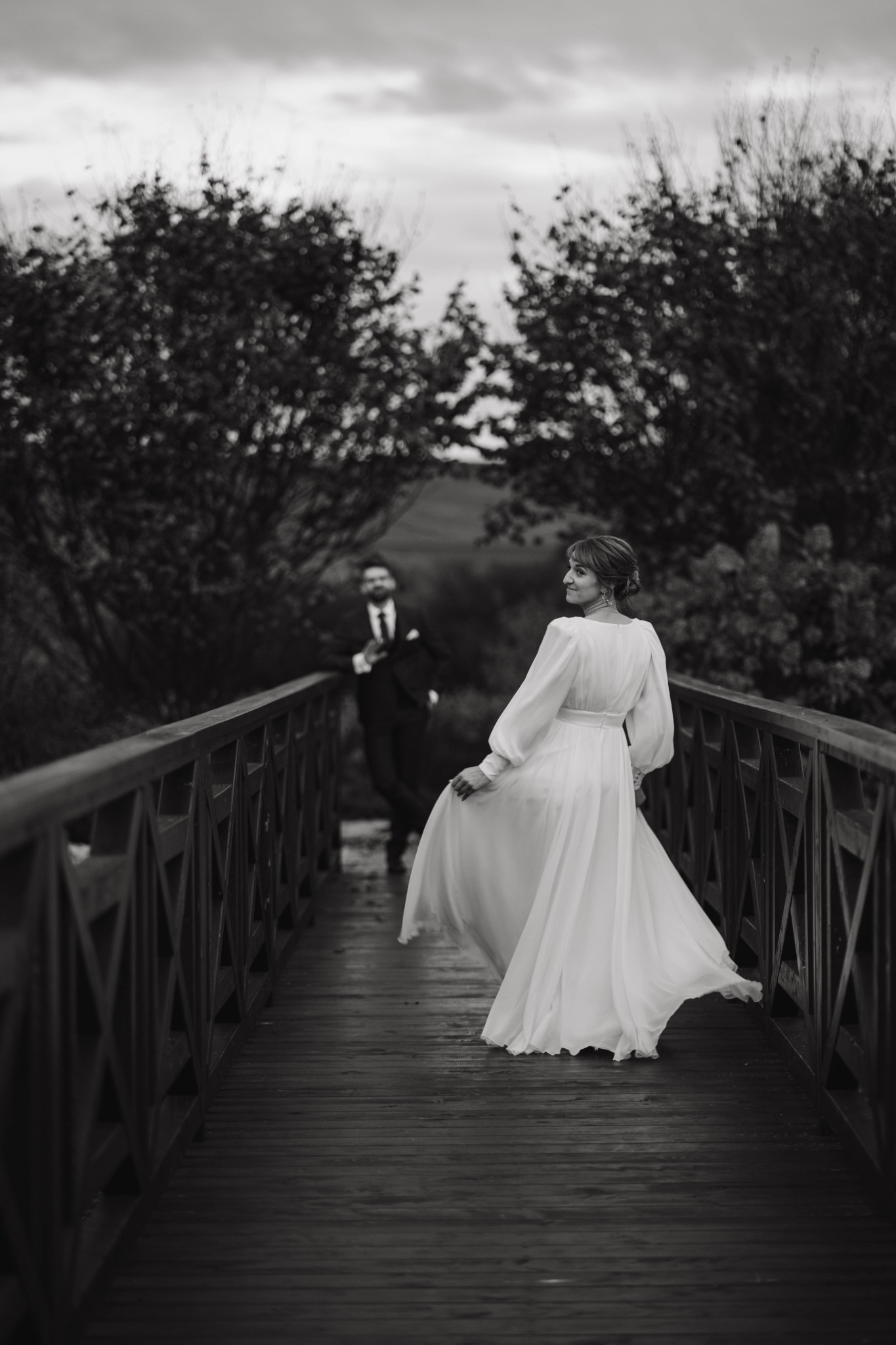 Czarno-białe zdjęcie panny młodej w zwiewnej sukni, odwracającej się na drewnianym moście, z panem młodym w tle, otoczone drzewami.