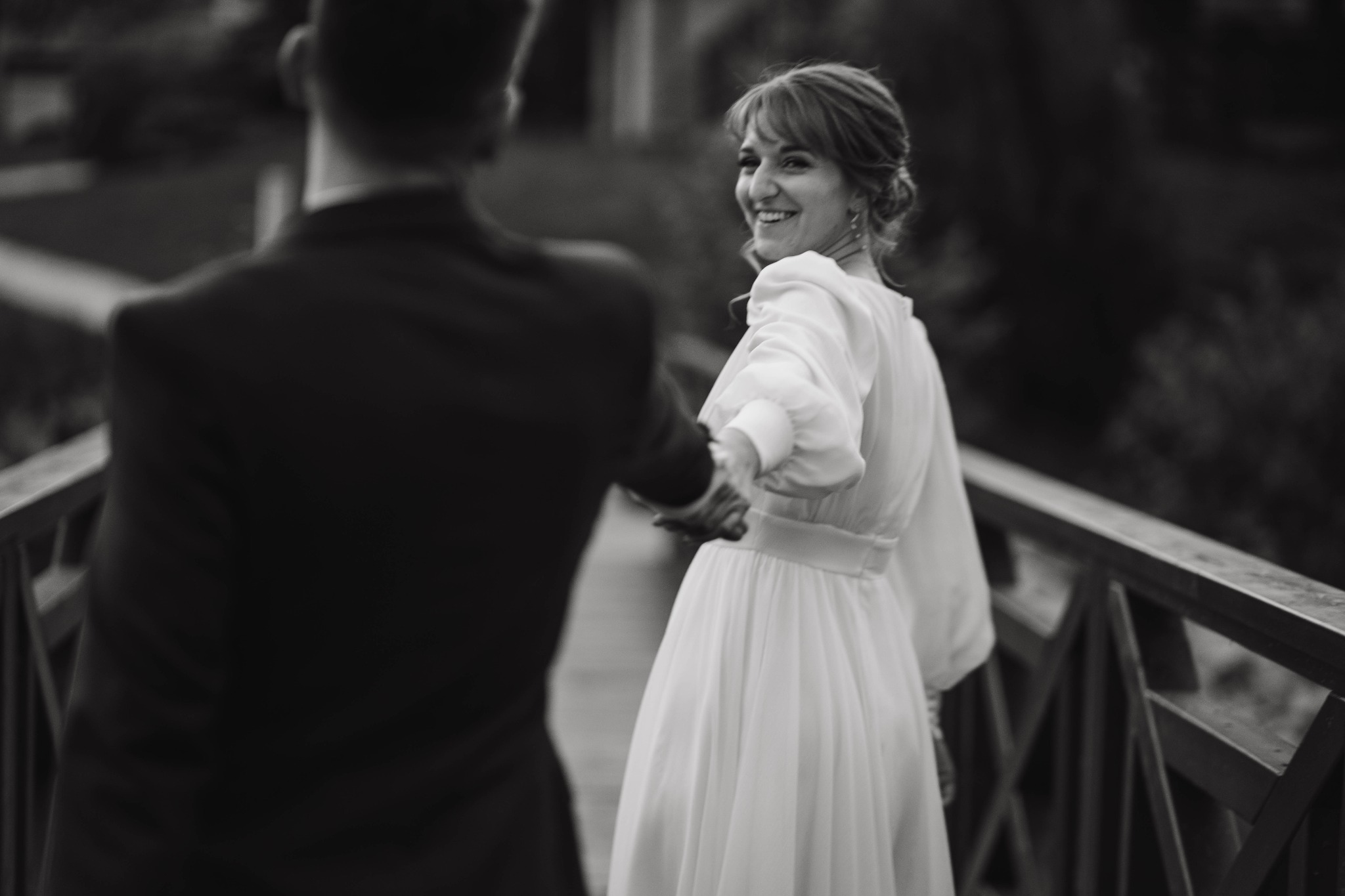 Czarno-białe zdjęcie panny młodej w zwiewnej sukni, odwracającej się z uśmiechem, trzymając za rękę pana młodego na drewnianym moście.