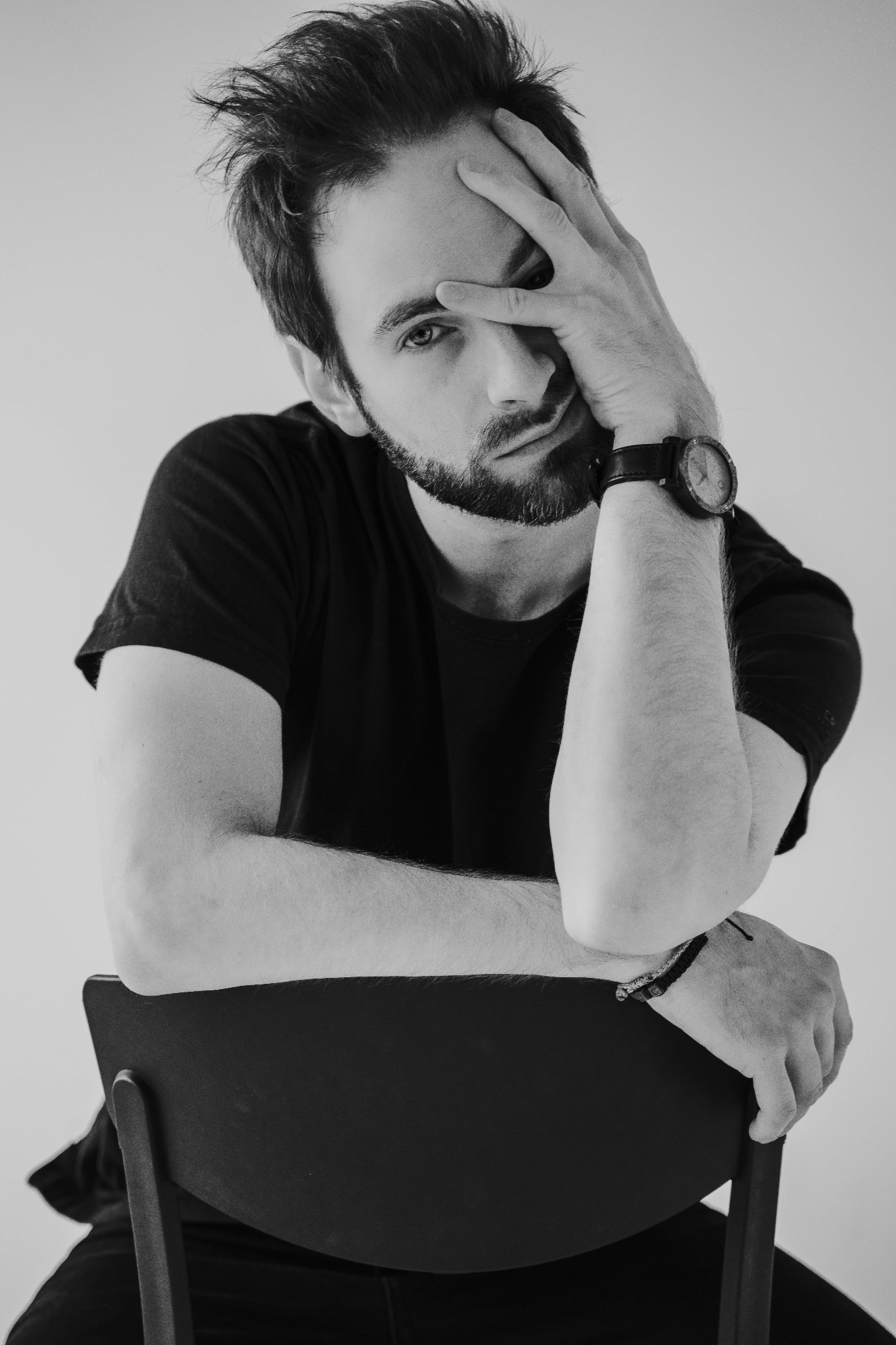Portret studyjny brodatego mężczyzny w czarnej koszulce, opierającego się o krzesło, z ręką na czole, w czarno-białej tonacji.