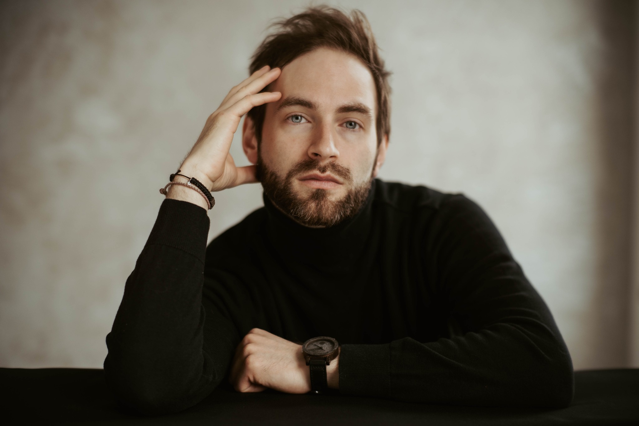 Portret mężczyzny z brodą w czarnym golfie, oparty zamyślony, z drewnianym zegarkiem na nadgarstku i bransoletkami z koralików.