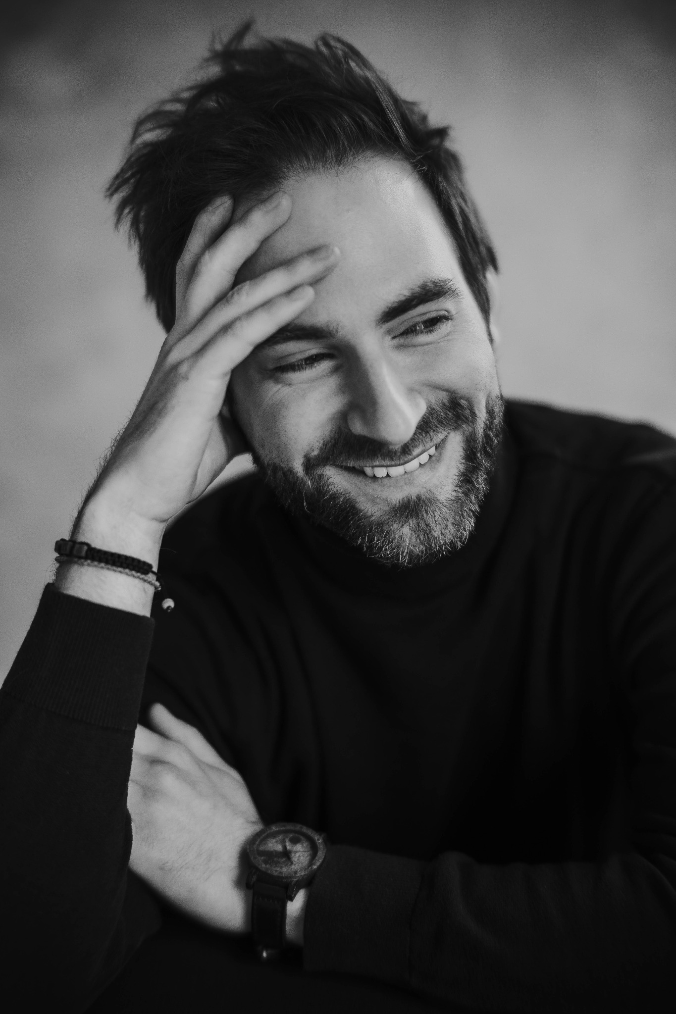 Czarno-biały portret uśmiechniętego mężczyzny z brodą, w czarnym golfie, opierającego głowę na dłoni z widocznym zegarkiem na nadgarstku i bransoletkami.