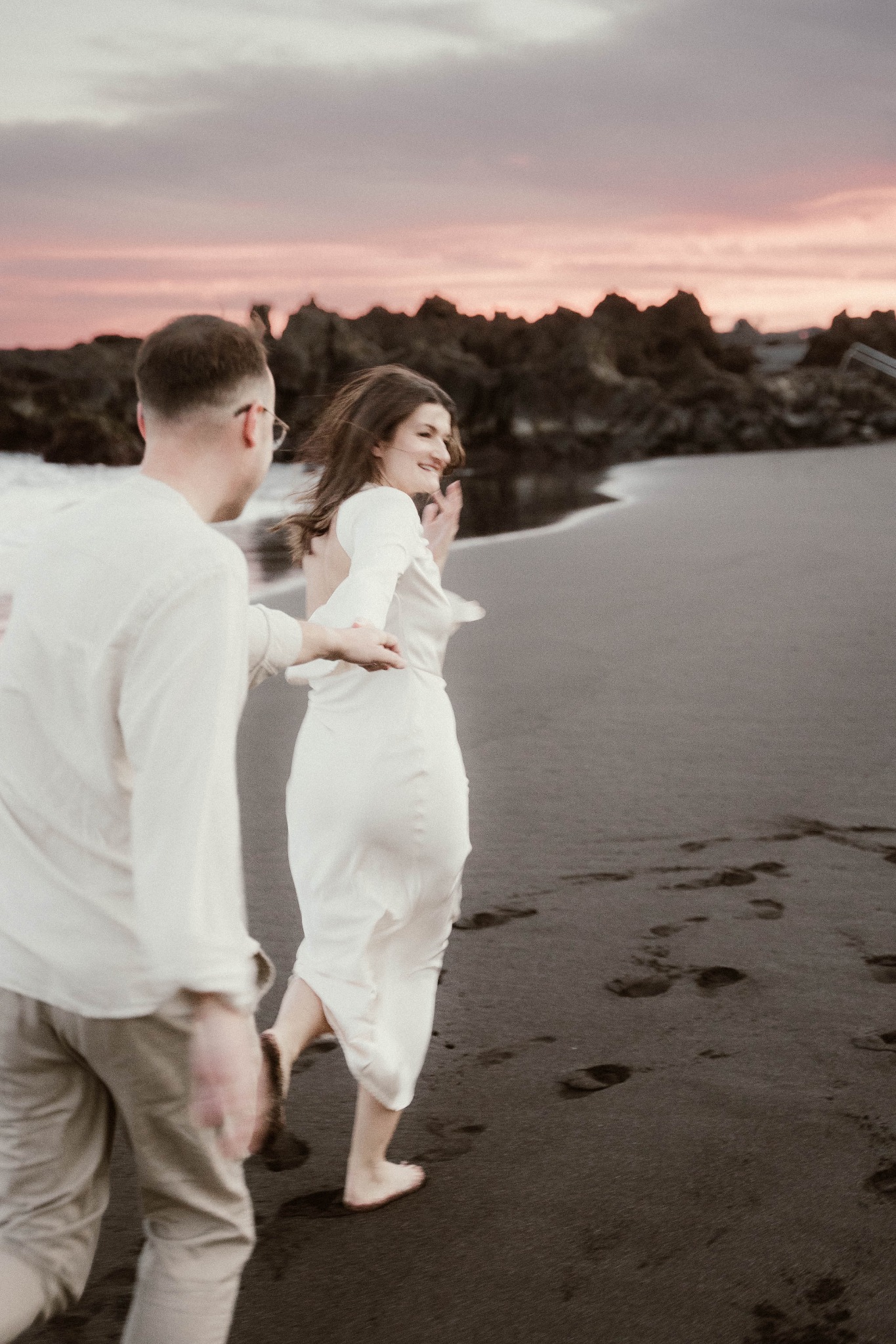 Para w jasnych ubraniach biegnie boso po czarnym piasku plaży, w tle widać skały i różowe niebo o zachodzie słońca, kobieta spogląda z uśmiechem przez ramię.