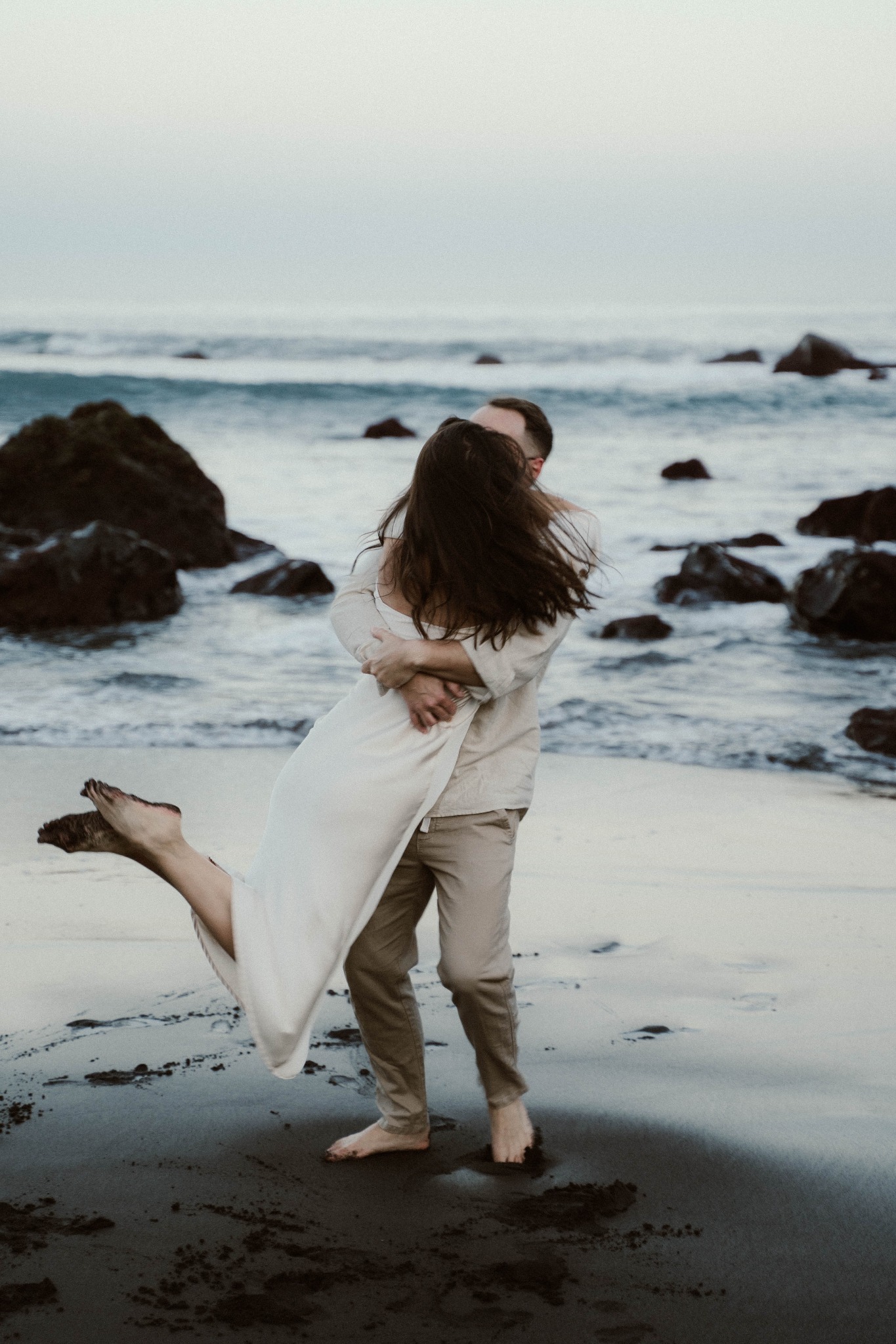 Para w objęciach na plaży; mężczyzna trzyma kobietę w białej sukience, w tle fale i skały.