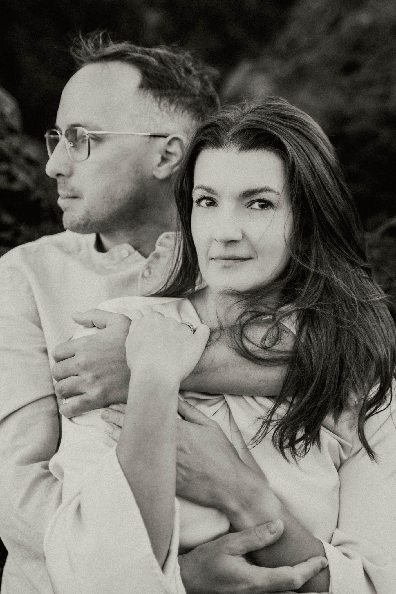 Czarno-białe portretowe ujęcie narzeczeńskie: mężczyzna w okularach obejmuje kobietę z pierścionkiem zaręczynowym na tle skalistego krajobrazu.