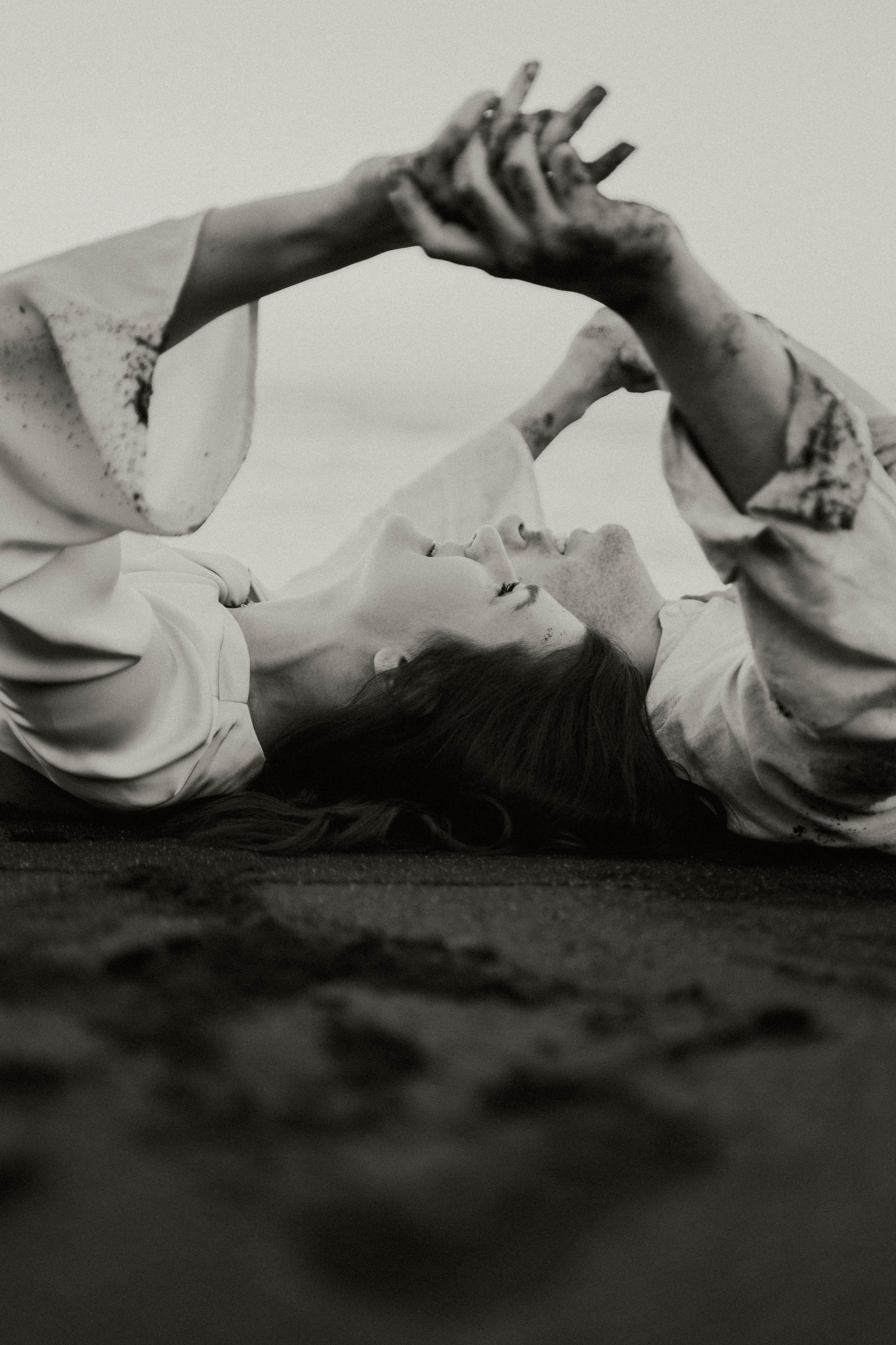 Czarno-białe zdjęcie pary leżącej na plaży, trzymającej się za ręce, z piaskiem na dłoniach i ubraniach, skierowanej twarzami ku sobie.