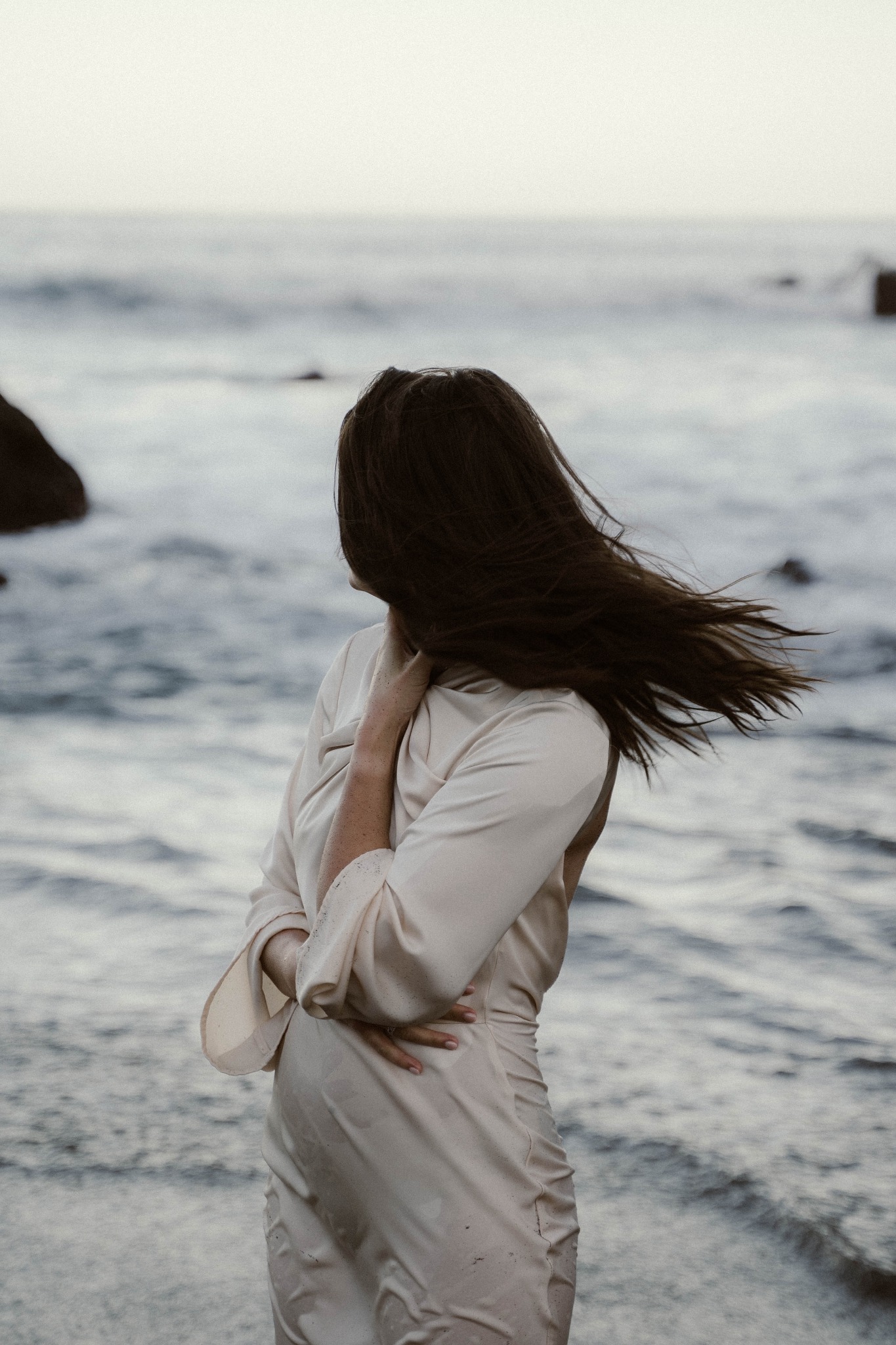 Kobieta w jasnej sukience stoi tyłem na plaży, włosy rozwiane na wietrze, w tle morze i skały.