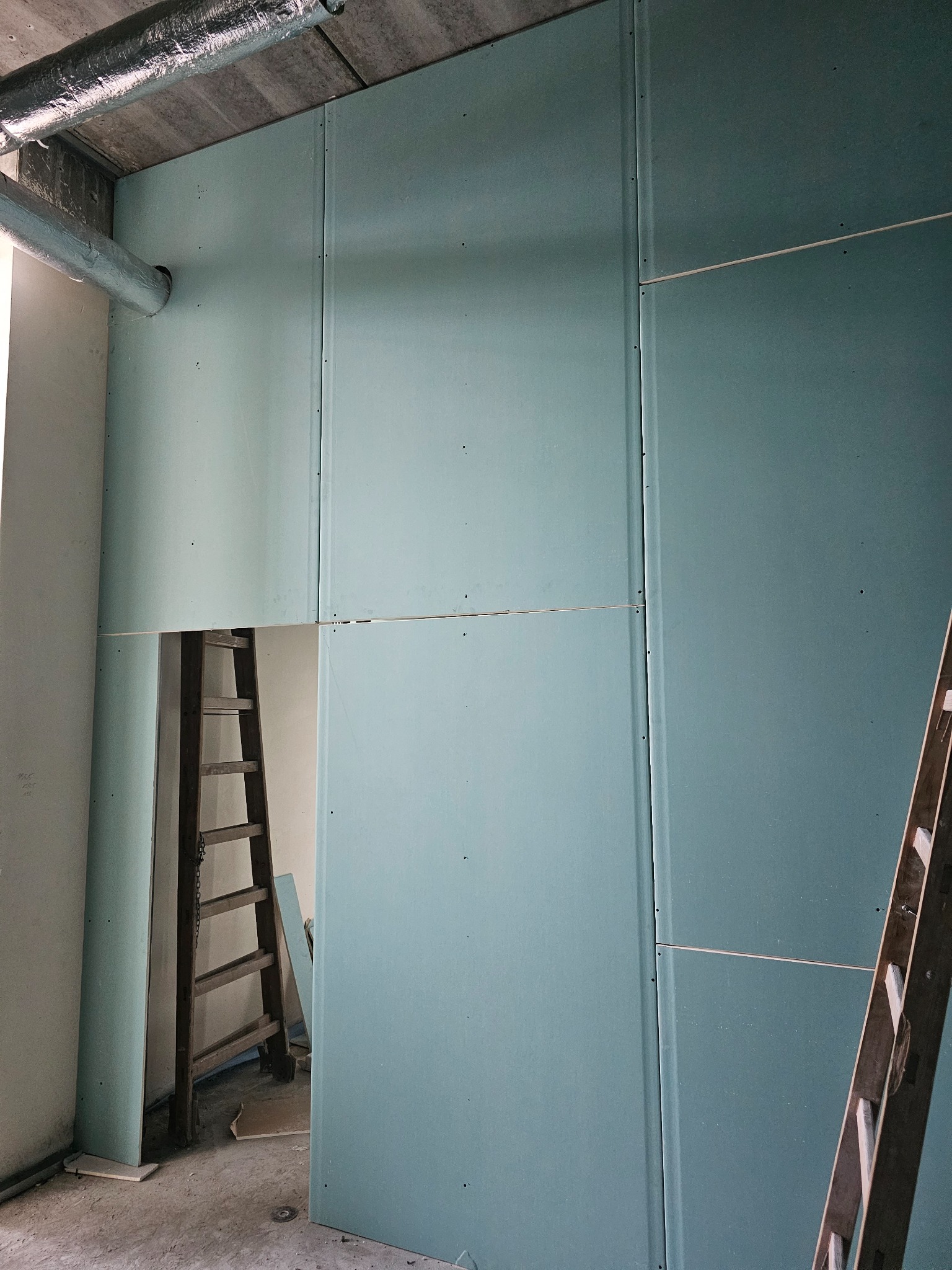 Ściana z płyt kartonowo-gipsowych w trakcie montażu, widoczne łączenia i drabiny pomocnicze, fragment instalacji wentylacyjnej pod sufitem.