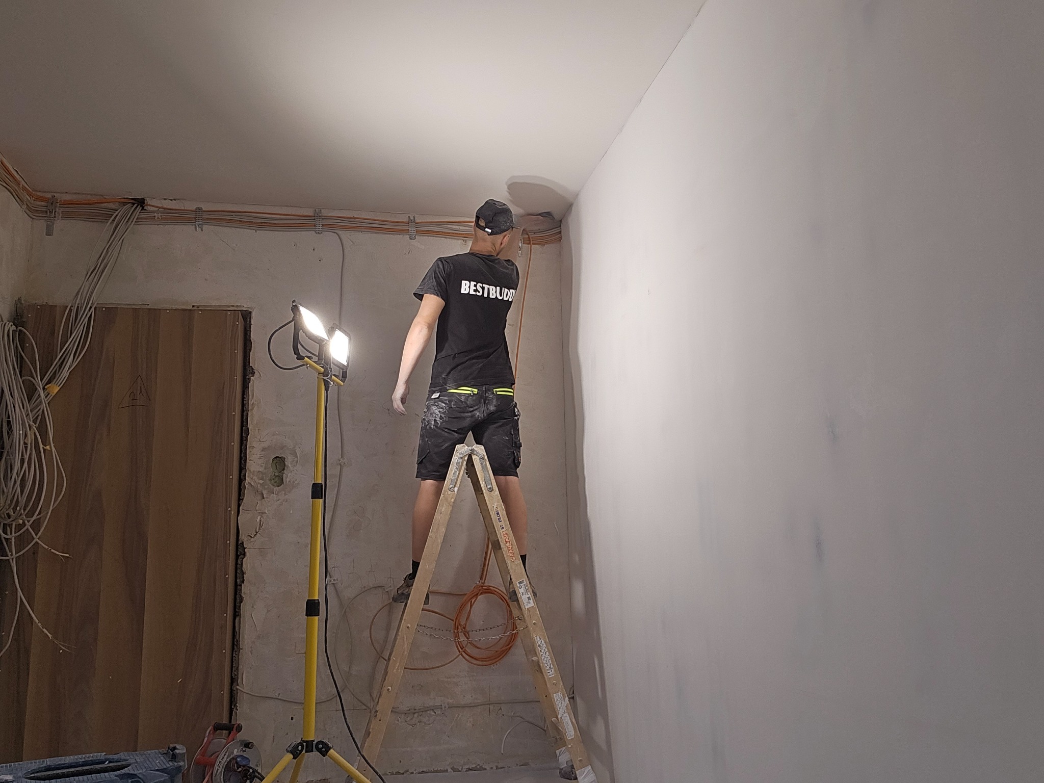 Pracownik na drabinie, w trakcie wyrównywania narożnika ściany w pomieszczeniu, widoczne przewody elektryczne pod sufitem i lampa halogenowa na statywie.