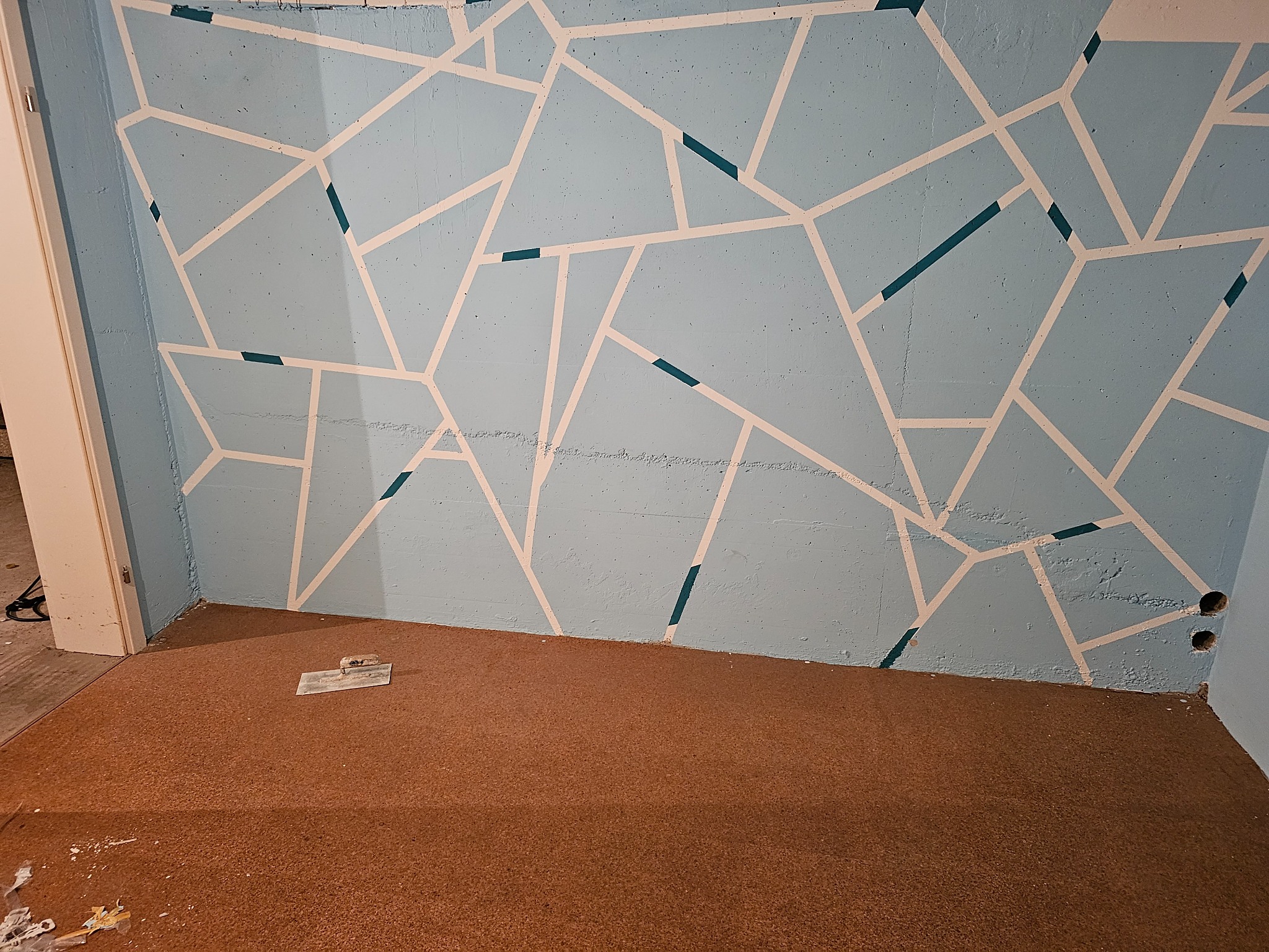Niebieska ściana z geometrycznym wzorem z białych linii, zabezpieczonych niebieską taśmą malarską, na podłodze brązowa wykładzina korkowa, widoczna szpachelka i fragment otwartych drzwi.
