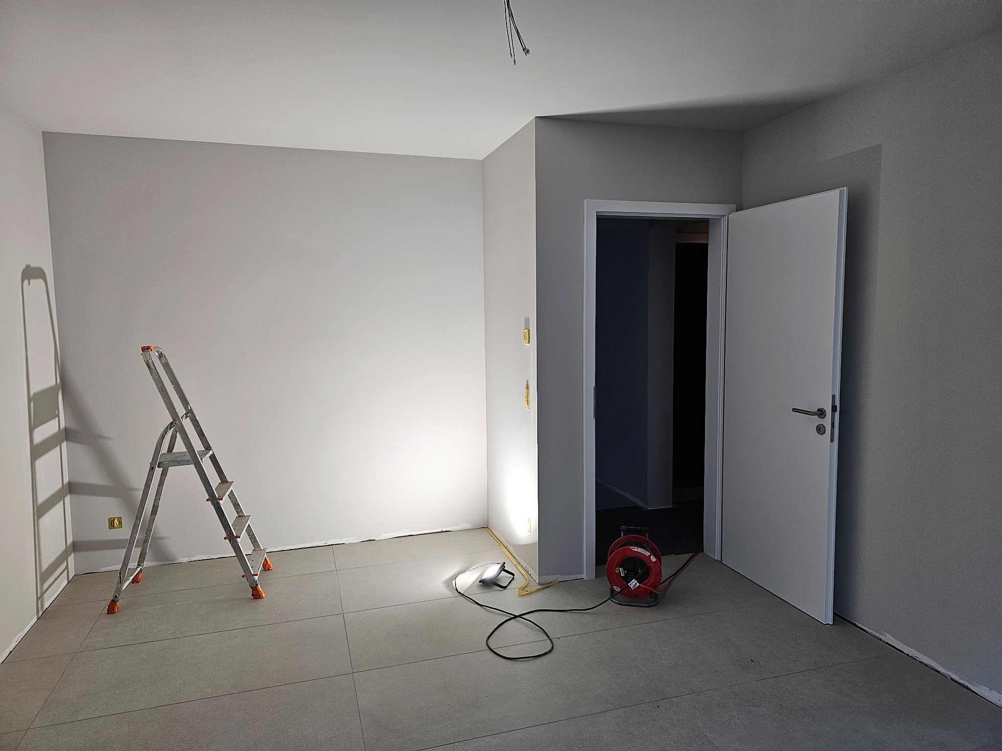 Pokój po świeżym pomalowaniu ścian na szaro, widoczna drabina aluminiowa, zwinięty przedłużacz na bębnie i halogen oświetlający ścianę przy podłodze, otwarte białe drzwi.