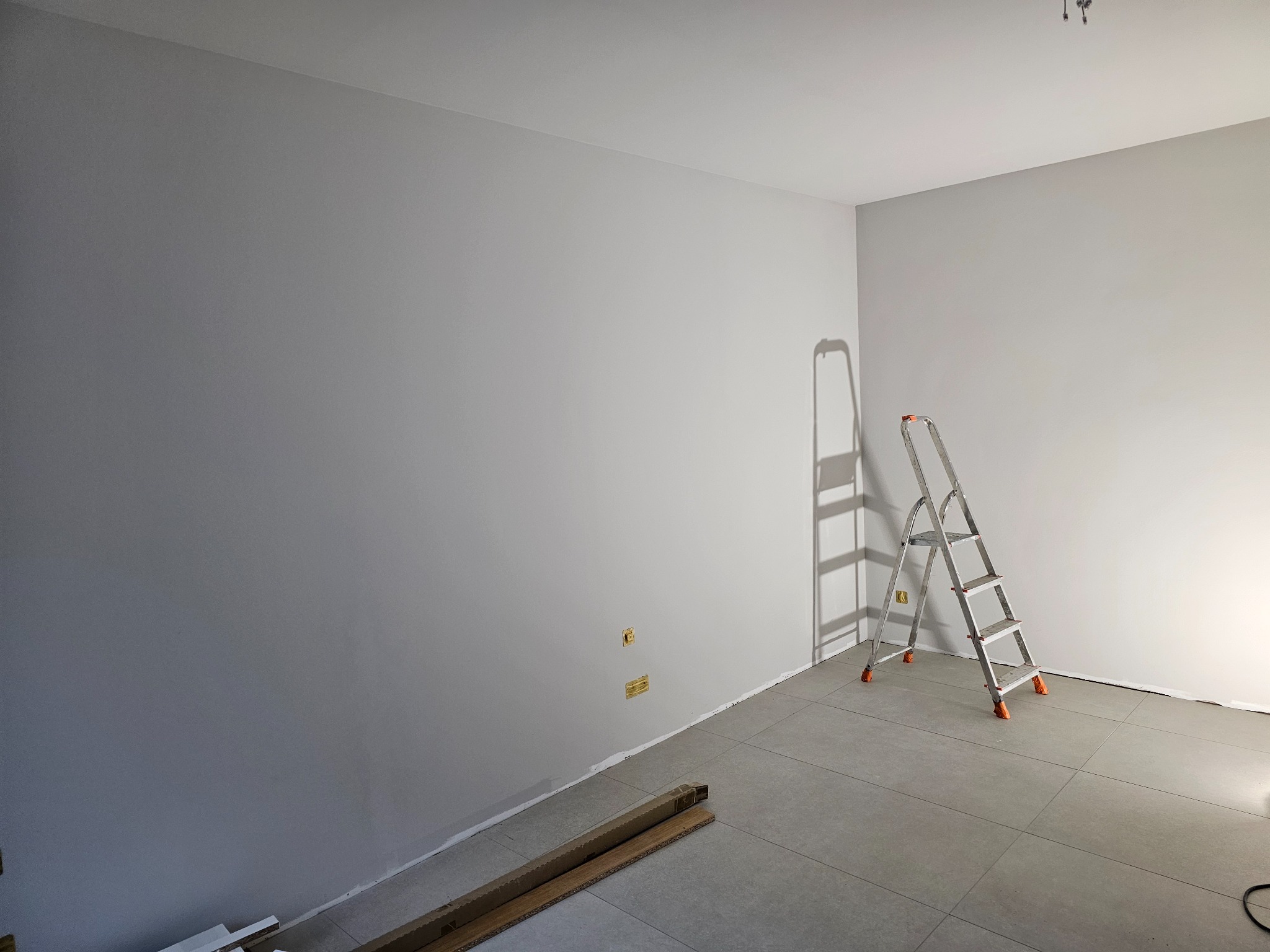 Wnętrze po świeżym pomalowaniu na szaro, widoczna aluminiowa drabina, kartony z listwami przypodłogowymi leżące na podłodze z płytek, gniazdka elektryczne na ścianie, ślady po taśmie malarskiej...