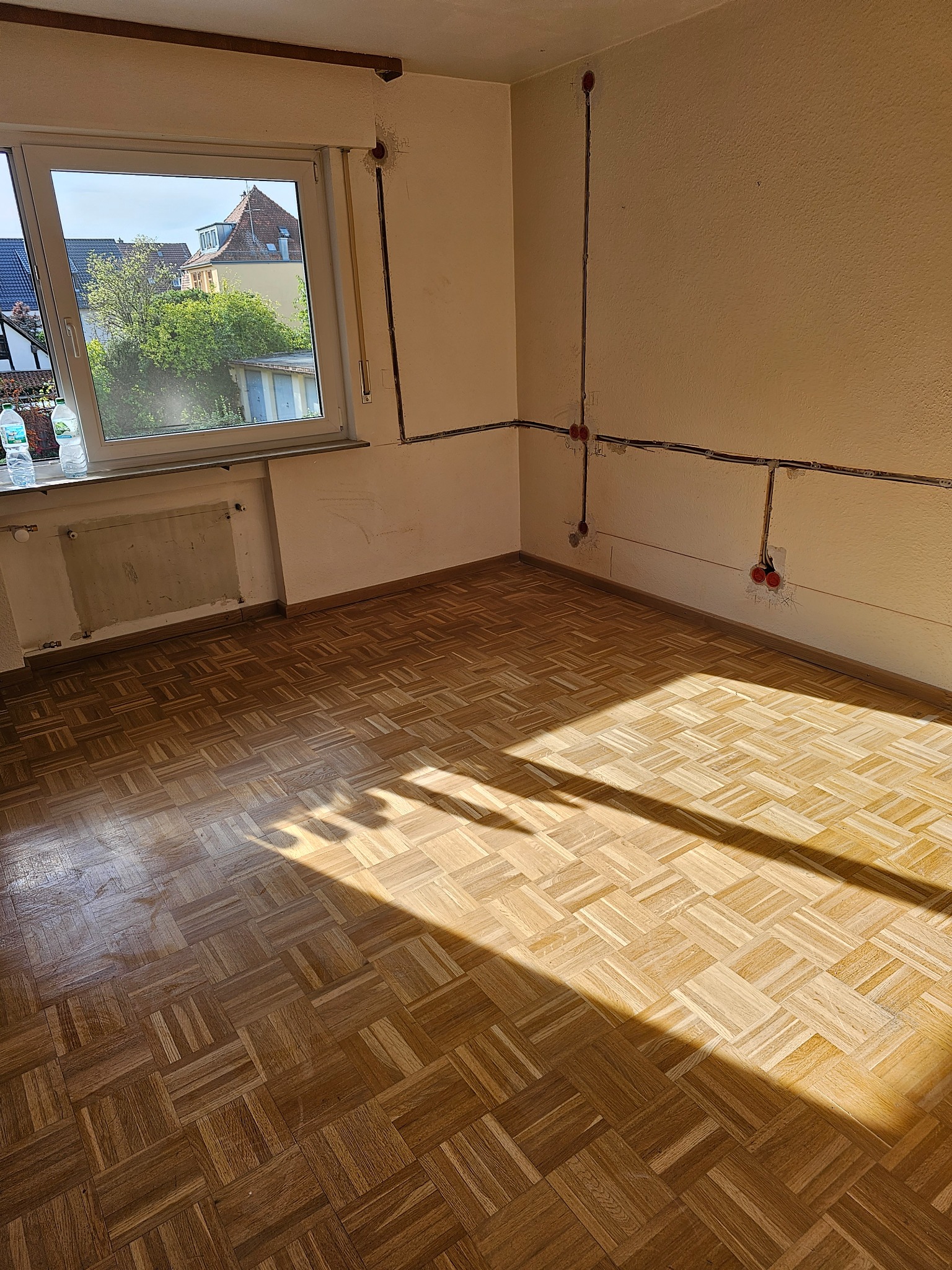 Pokój w trakcie remontu: parkiet w jodełkę, odsłonięte przewody elektryczne na ścianach, widok z okna na budynki i zieleń.