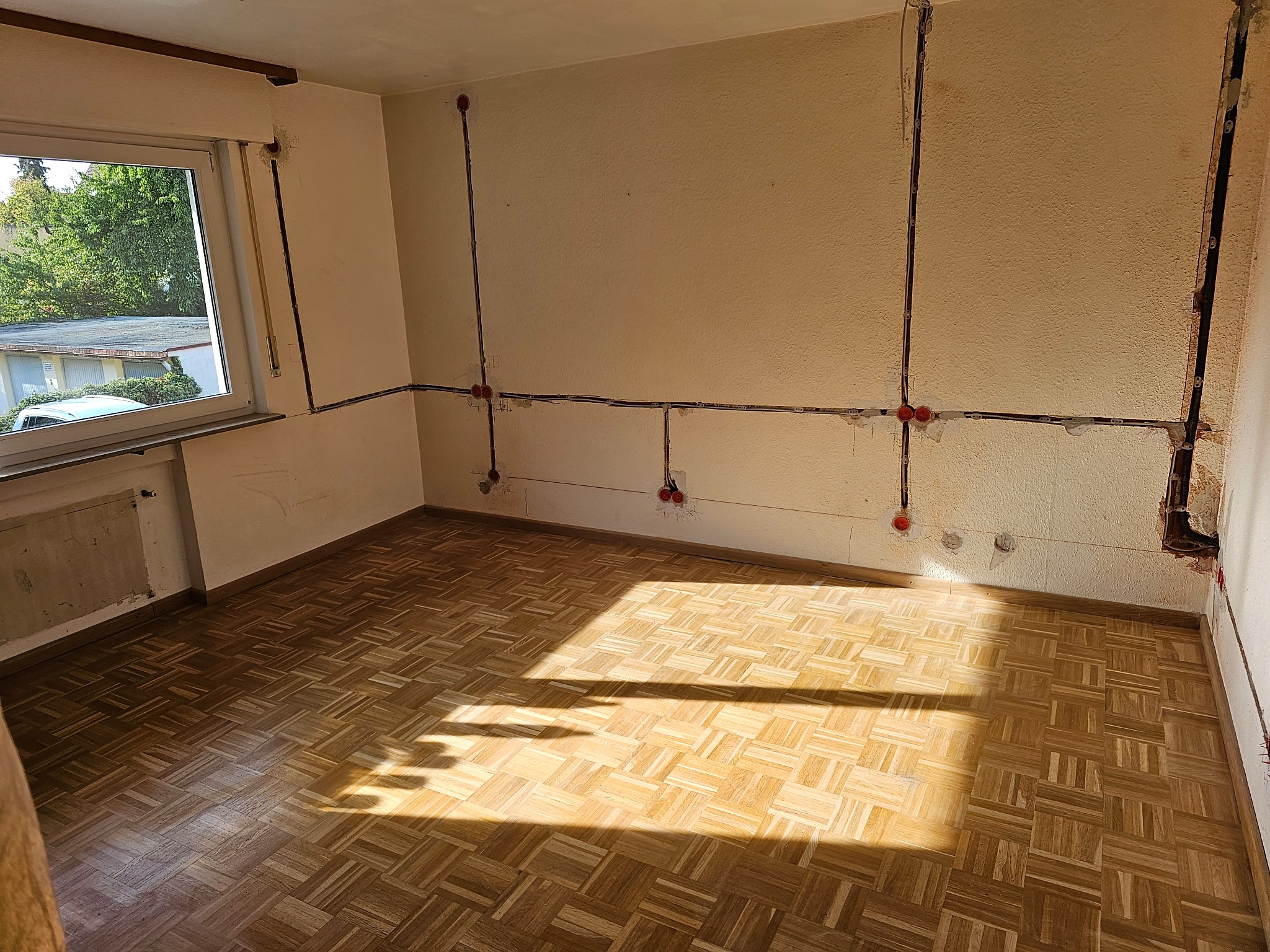 Pokój w trakcie remontu: odsłonięte przewody elektryczne w ścianach, przygotowane do instalacji gniazdek, parkiet w jodełkę oświetlony promieniami słońca wpadającymi przez okno.