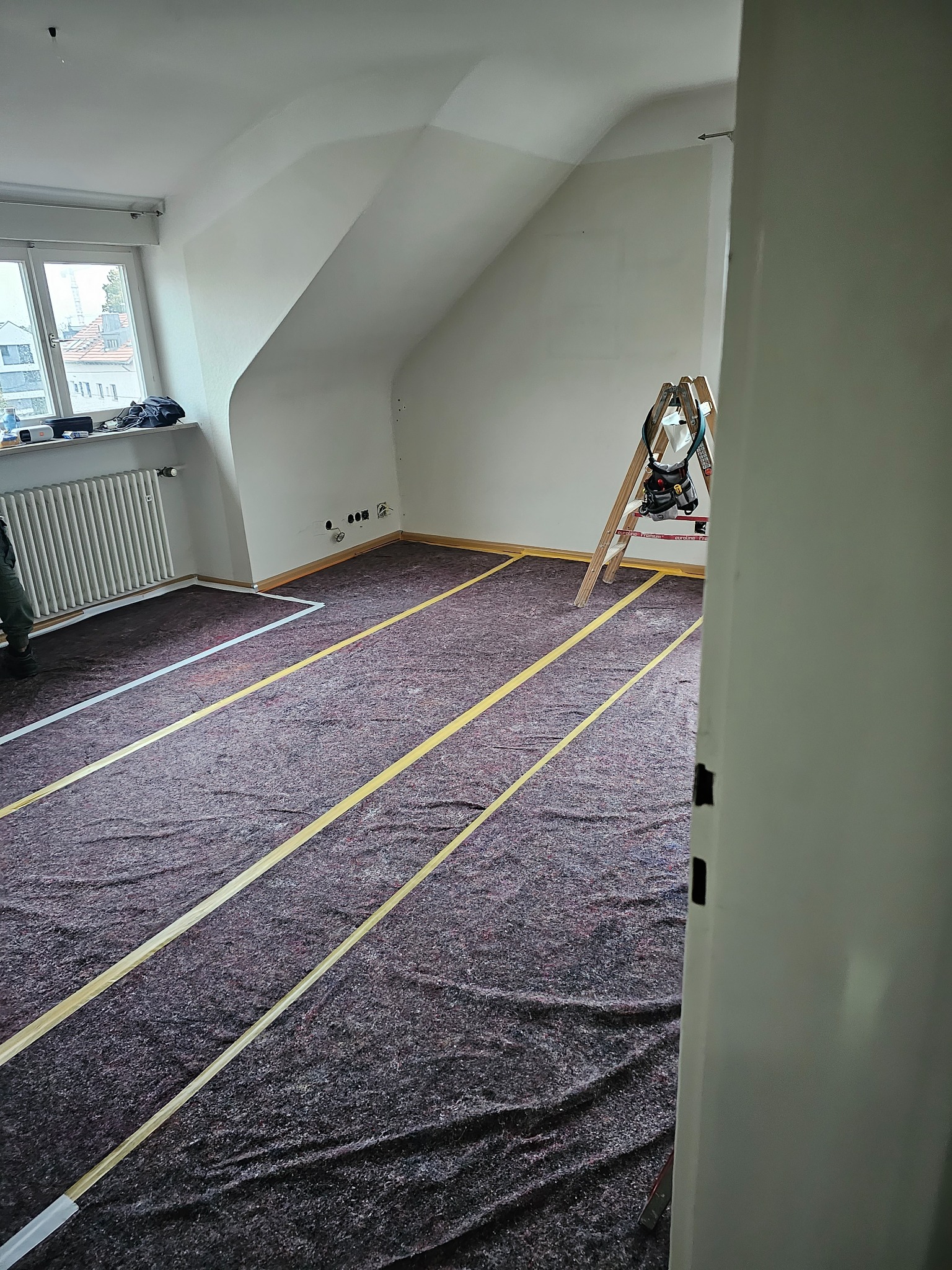 Pokój na poddaszu w trakcie zabezpieczania podłoża przed pracami wykończeniowymi; podłoga wyłożona grubą włókniną ochronną z taśmami wyznaczającymi obszar roboczy, drabina z narzędziami oparta...