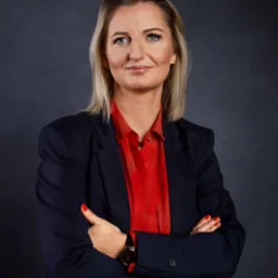 Portret kobiety w granatowej marynarce i czerwonej koszuli, z założonymi rękami, patrzącej w obiektyw, z delikatnym uśmiechem, na ciemnoszarym tle.