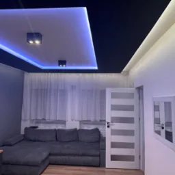 Nowoczesny salon z podwieszanym sufitem z oświetleniem LED, szary narożnik i białe drzwi z poziomymi przeszkleniami.