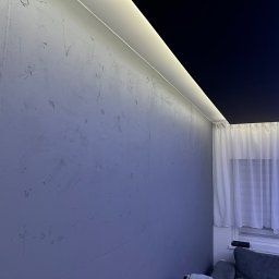 Decor Luk - Ściana z szarą, nierówną fakturą, oświetlona listwą LED wzdłuż sufitu, z widocznym fragmentem czarnego sufitu podwieszanego i firanką w oknie.