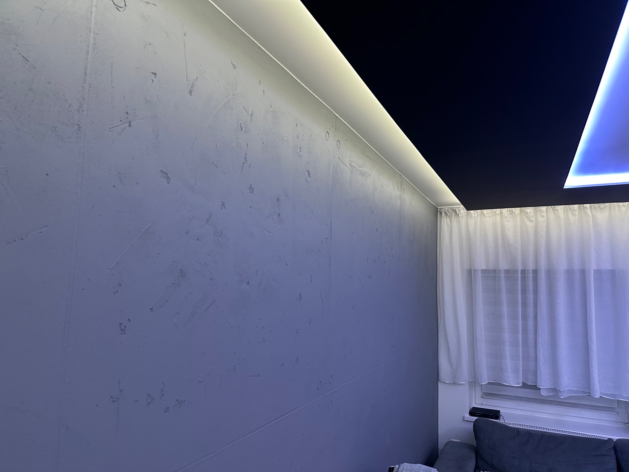 Ściana z szarą, nierówną fakturą, oświetlona listwą LED wzdłuż sufitu, z widocznym fragmentem czarnego sufitu podwieszanego i firanką w oknie.
