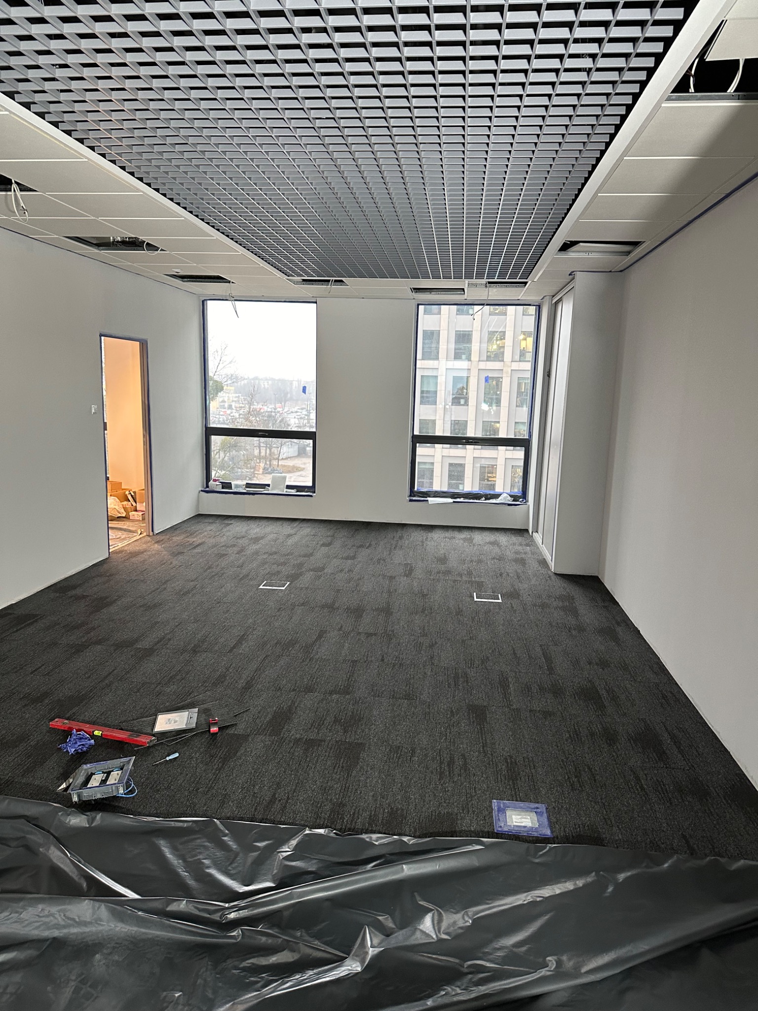 Biuro w trakcie wykańczania: szara wykładzina dywanowa, podwieszany sufit rastrowy, dwa okna z widokiem na miasto, narzędzia na podłodze przykryte folią ochronną.