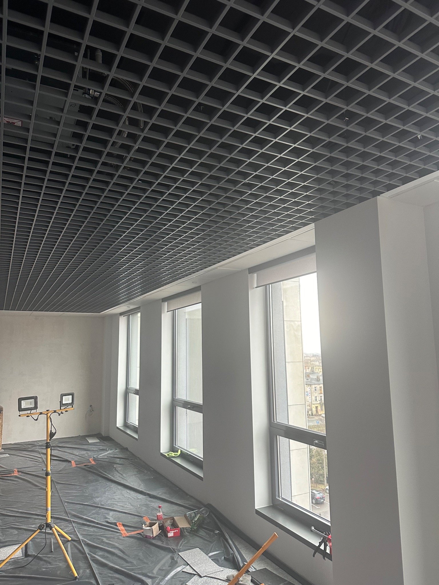 Sufit rastrowy w trakcie montażu w jasnym pomieszczeniu z trzema oknami, podłoga zabezpieczona folią, widoczny statyw z lampami halogenowymi.