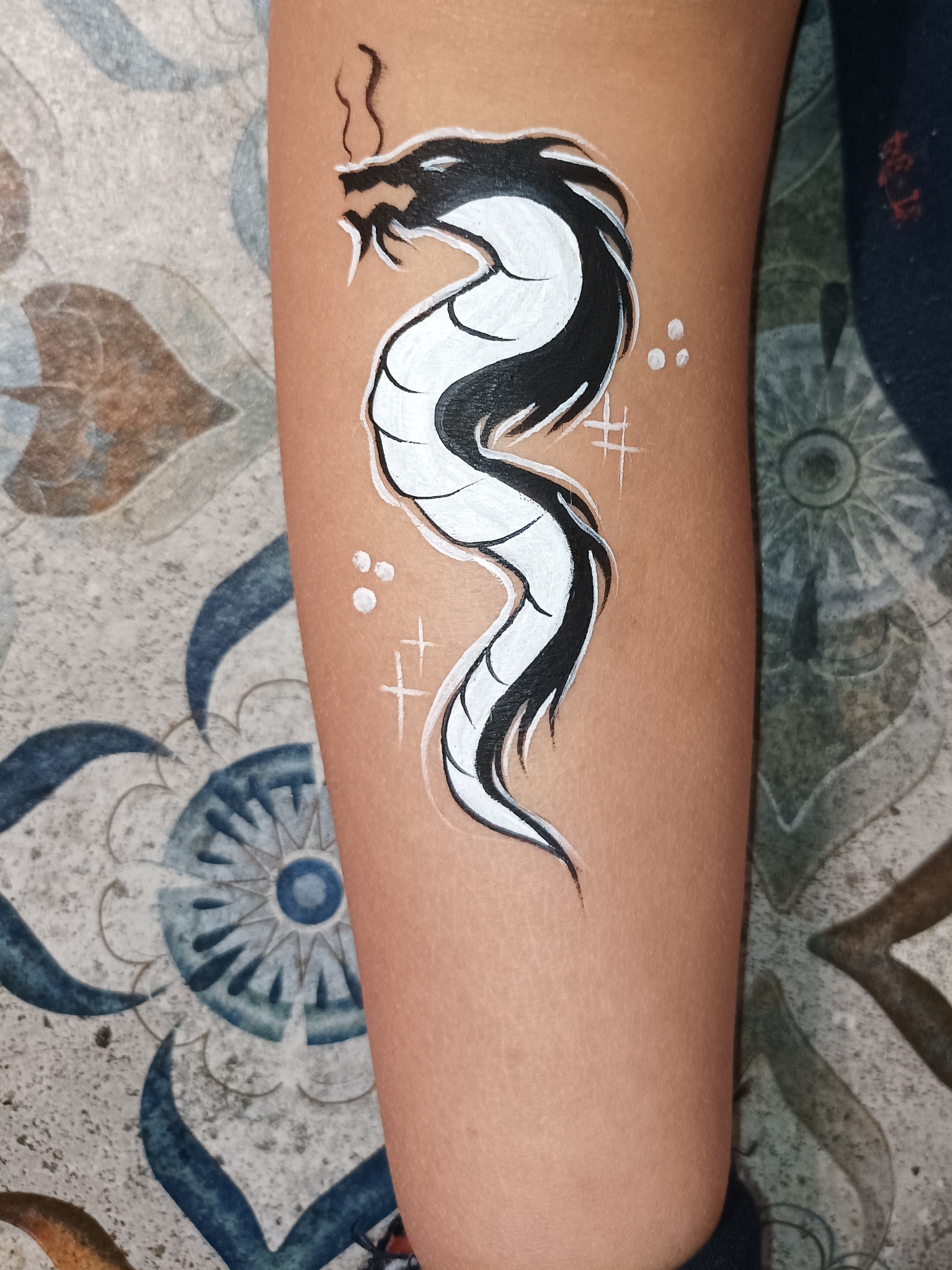 Ręka dziecka z namalowanym czarno-białym smokiem w stylu body painting, uzupełnionym o białe kropki i krzyżyki.