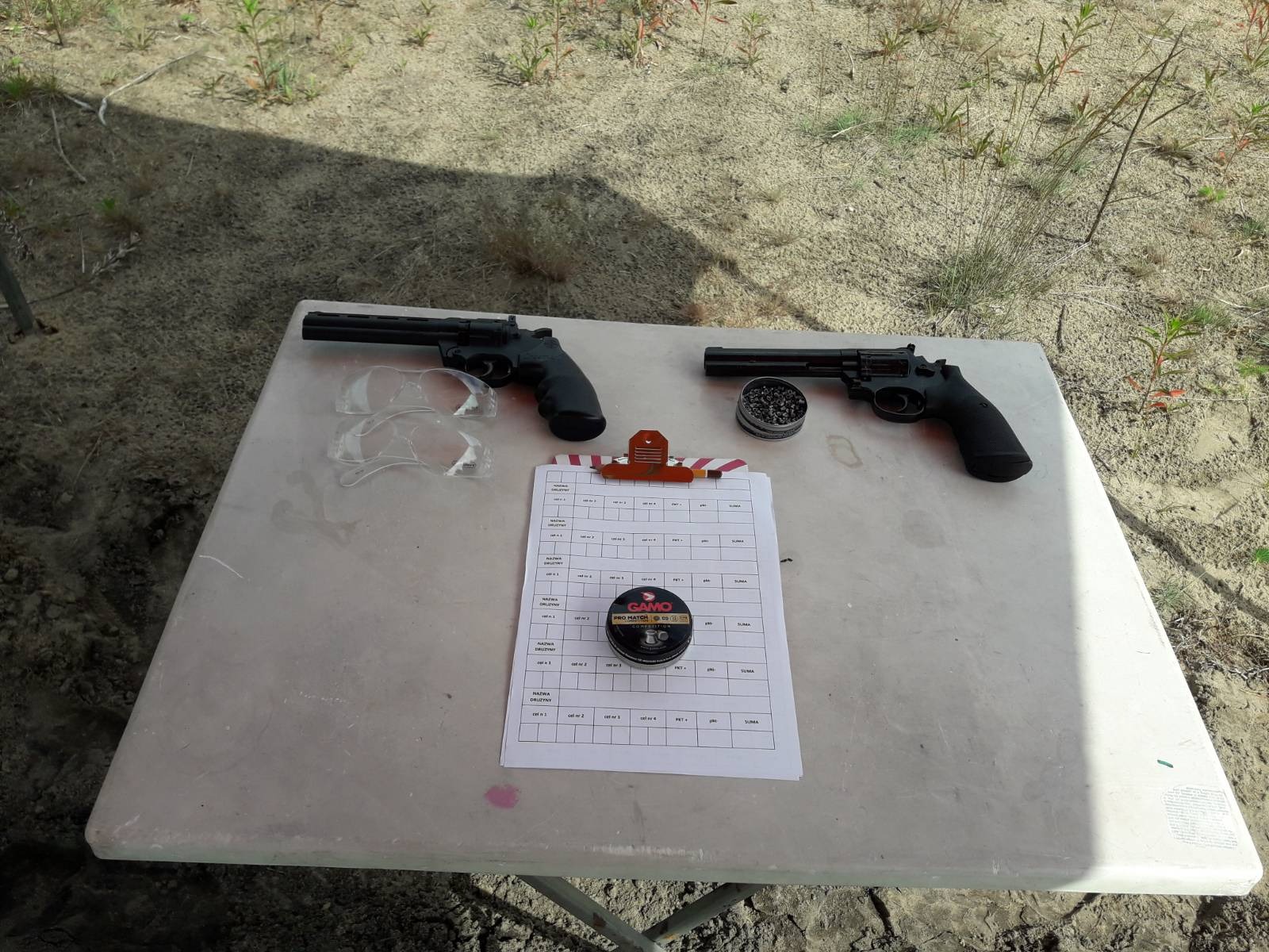 Dwa rewolwery pneumatyczne, okulary ochronne, tarcza strzelecka z klipsem i puszka z amunicją na białym stole na tle piaszczystego terenu z roślinnością.