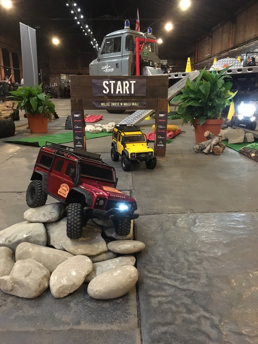 Miniaturowy tor przeszkód z modelami Land Rover Defender: czerwony pokonuje kamienie, żółty czeka za bramą startową, a szary na lawecie w tle, wszystko w hali z oświetleniem.