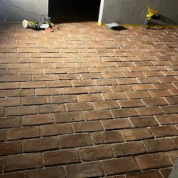 Ukończona podłoga z cegieł klinkierowych ułożonych w jodełkę, widoczne narzędzia pracy i lampa oświetleniowa, ściana z szarą strukturą.