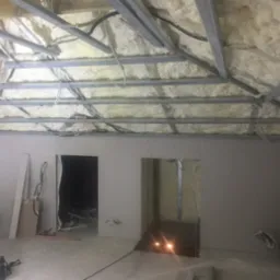 Poddasze w trakcie adaptacji: ocieplenie wełną, metalowa konstrukcja sufitu, ściany obłożone płytami kartonowo-gipsowymi z wyciętymi otworami na drzwi, widoczne instalacje.