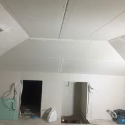 Montaż płyt karton gips na skośnym poddaszu z widocznymi otworami na drzwi, niezaszpachlowane łączenia i narzędzia budowlane w rogu.