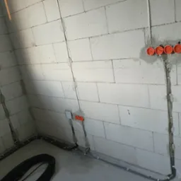 Surowa ściana z betonu komórkowego z widocznymi puszkami elektrycznymi, przewodami w peszlach i kablem na podłodze w trakcie prac.