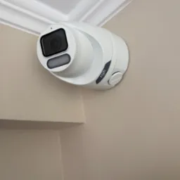 Kamera monitoringu UNV zamontowana w rogu pomieszczenia, widoczna na tle jasnej ściany i białego sufitu z ozdobnym gzymsem.