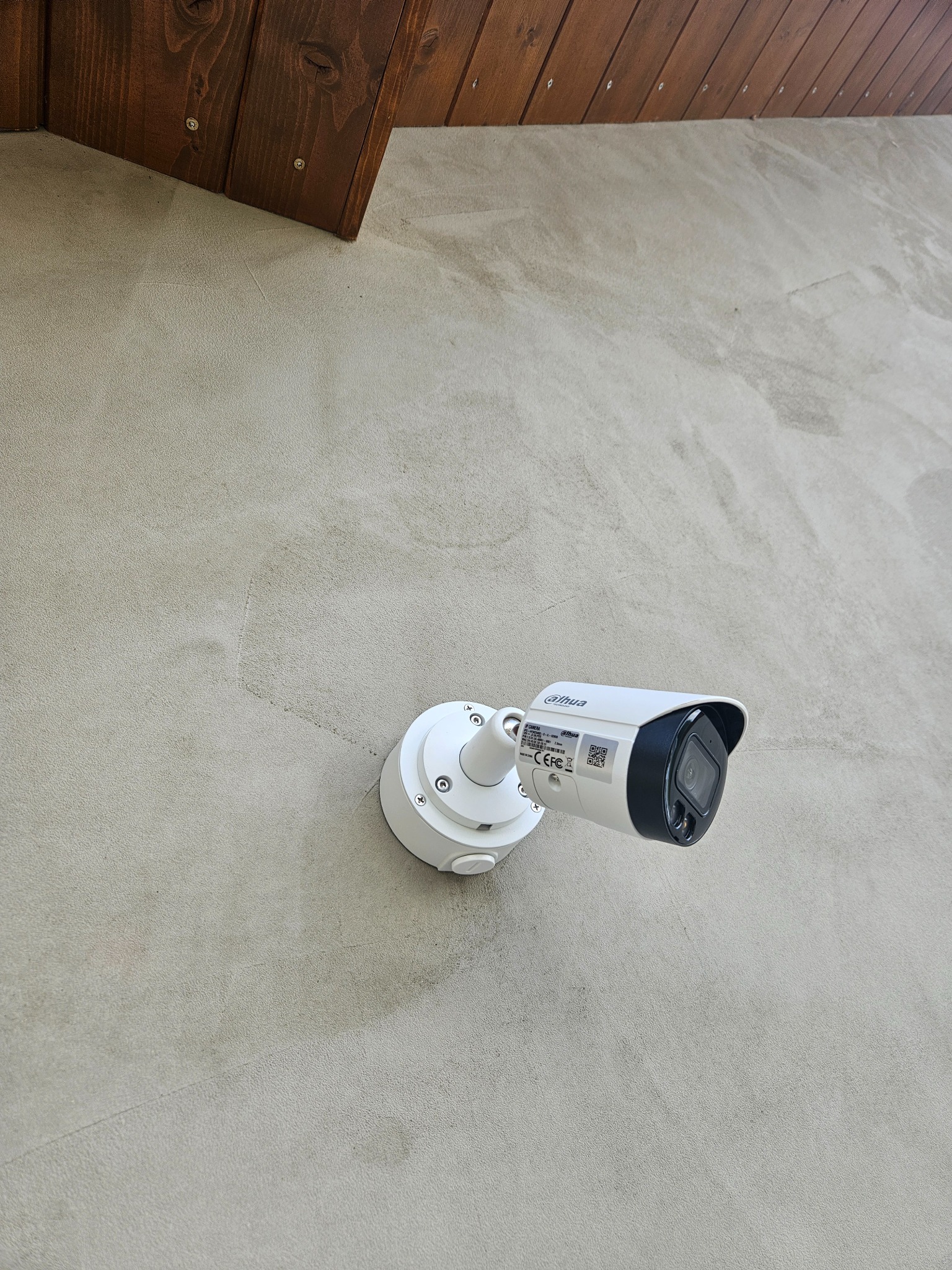 Biała kamera monitoringu Dahua zamontowana na jasnoszarej, teksturowanej ścianie pod drewnianym okapem.