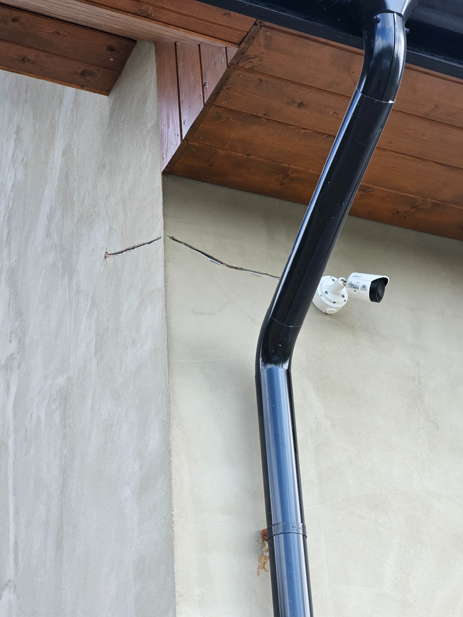 Kamera monitoringu przymocowana do ściany budynku obok rynny spustowej, widoczne niedociągnięcia w elewacji i wystające przewody.