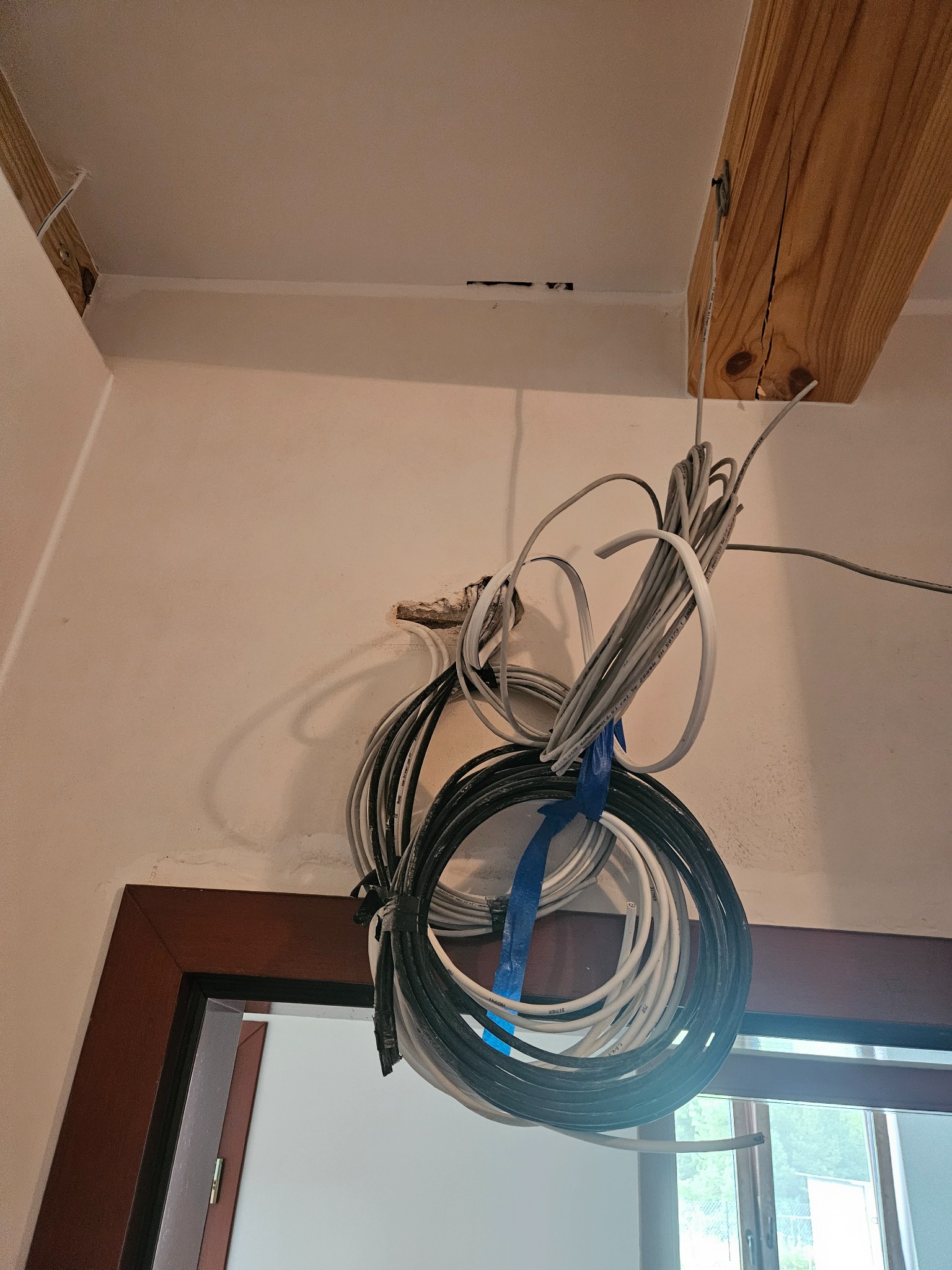 Wiązka kabli elektrycznych i telekomunikacyjnych zwisająca z sufitu, częściowo przymocowana do drewnianej belki, widoczne niedokończone prace instalacyjne.