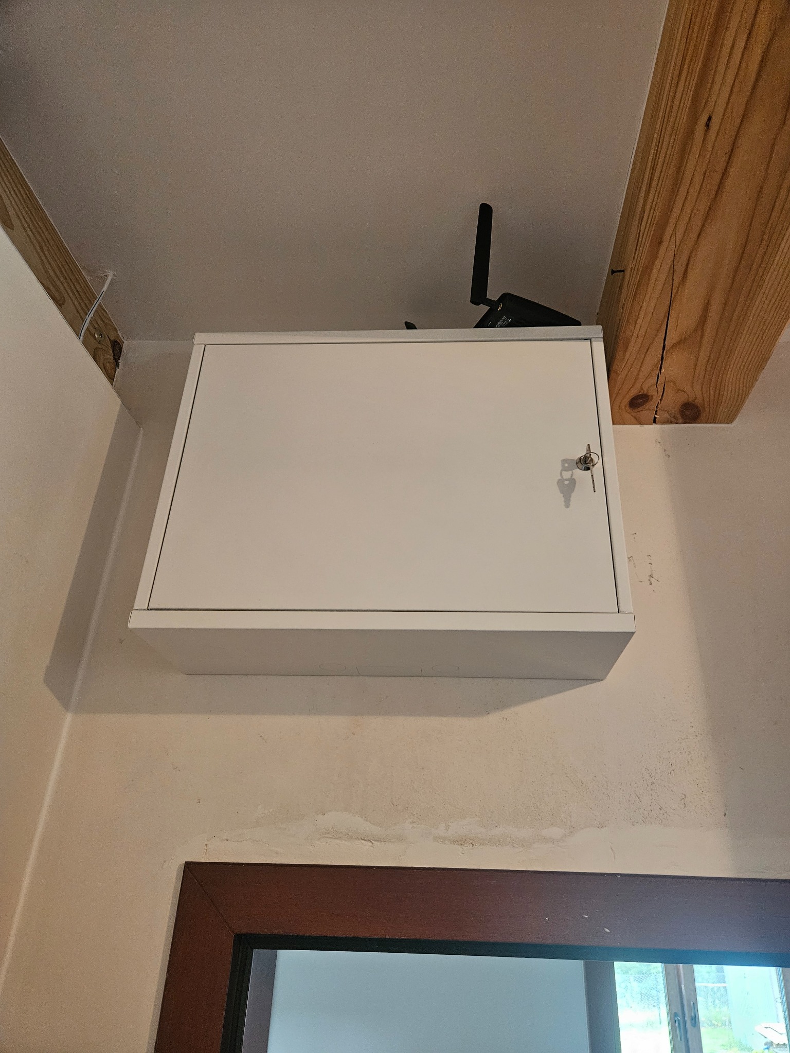 Biała skrzynka z kluczem wystającym z zamka, zamontowana na ścianie pod sufitem, obok drewnianej belki stropowej. Na górze skrzynki widoczny router z antenką.