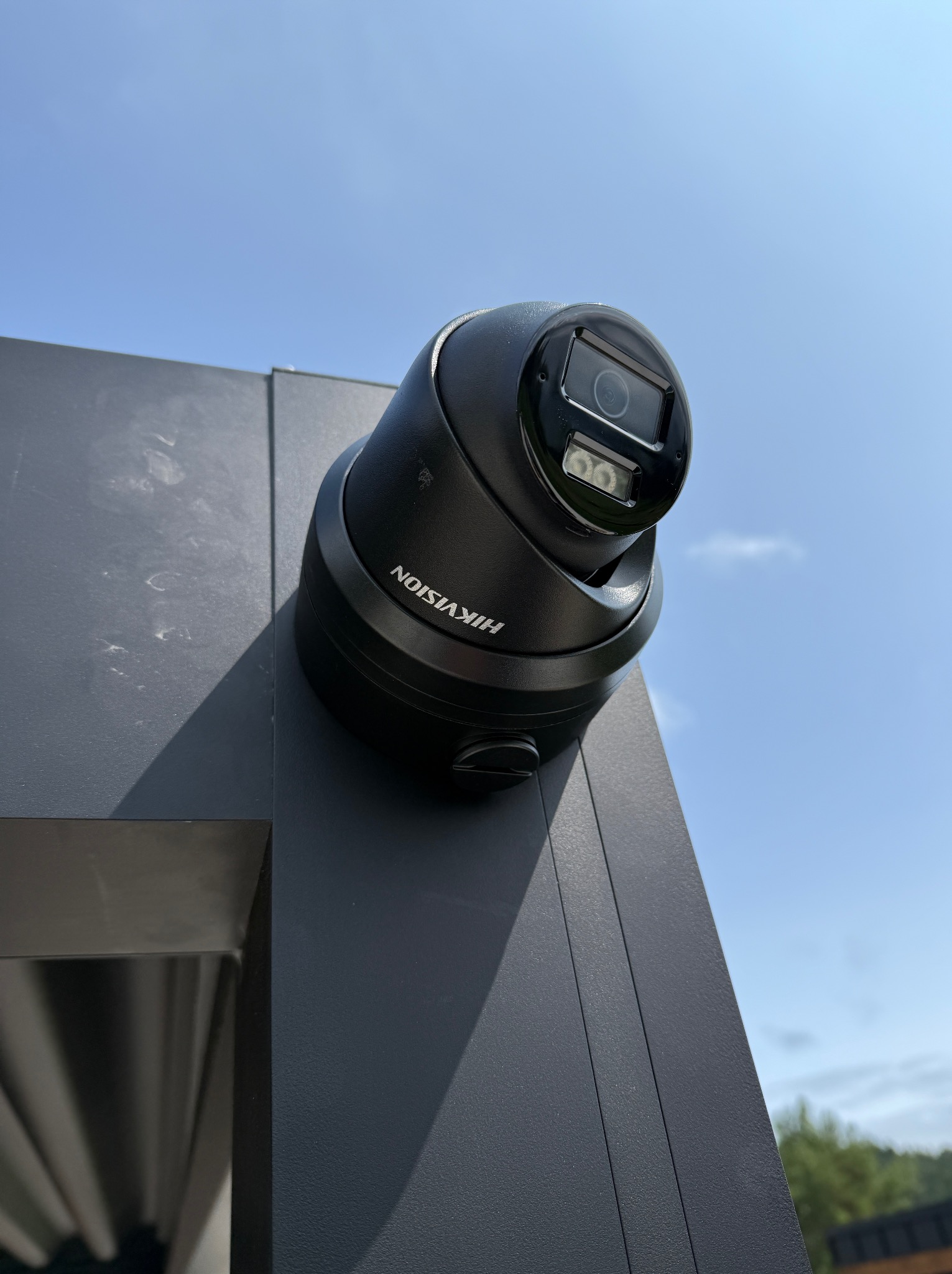 Kamera Hikvision typu dome, zamontowana na grafitowej elewacji budynku, skierowana w górę, na tle błękitnego nieba. Widok z dołu podkreśla solidność instalacji.