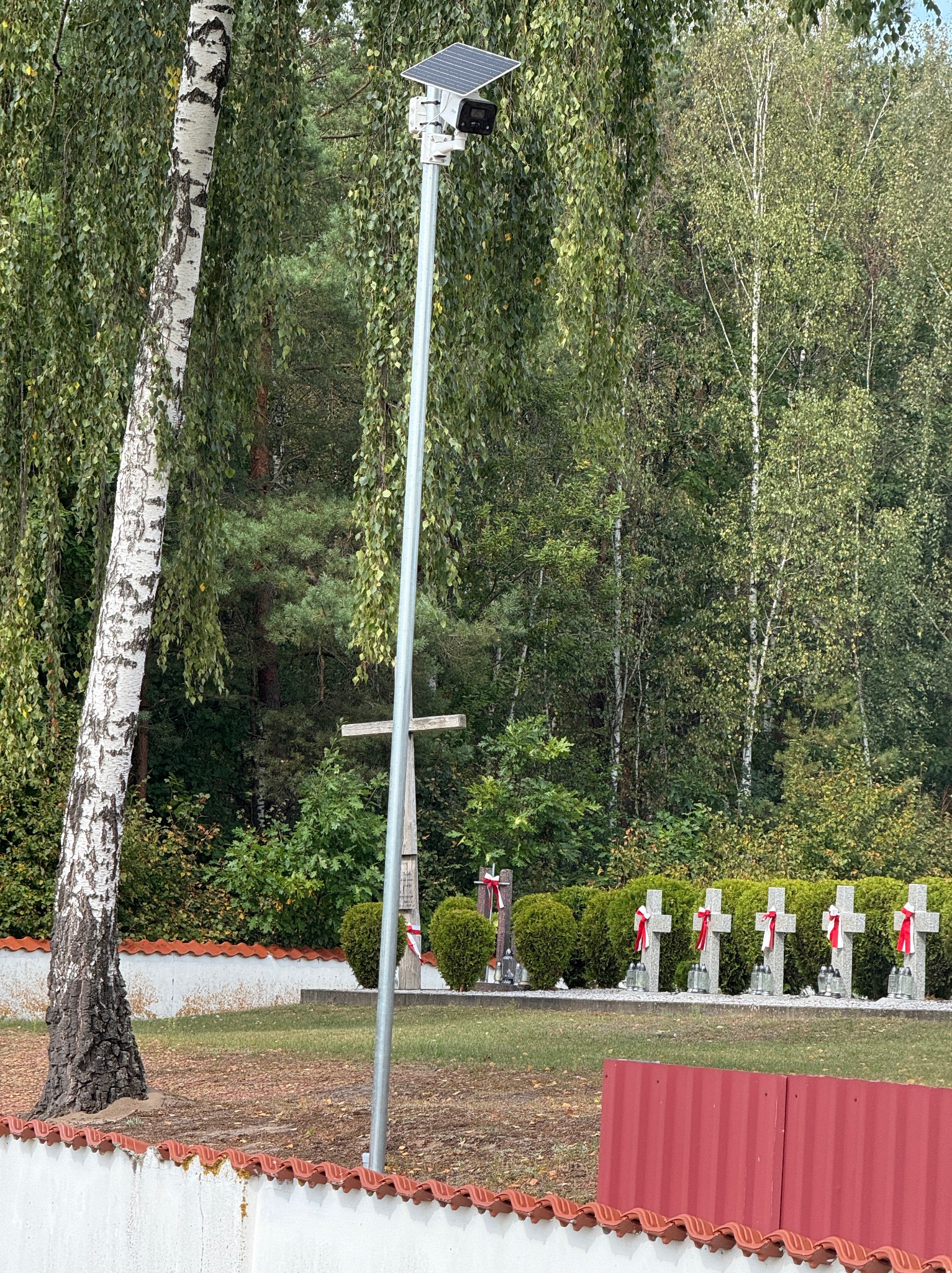 Kamera monitoringu na słupie z panelem solarnym, widok na cmentarz z krzyżami ozdobionymi biało-czerwonymi wstążkami, drzewa w tle.