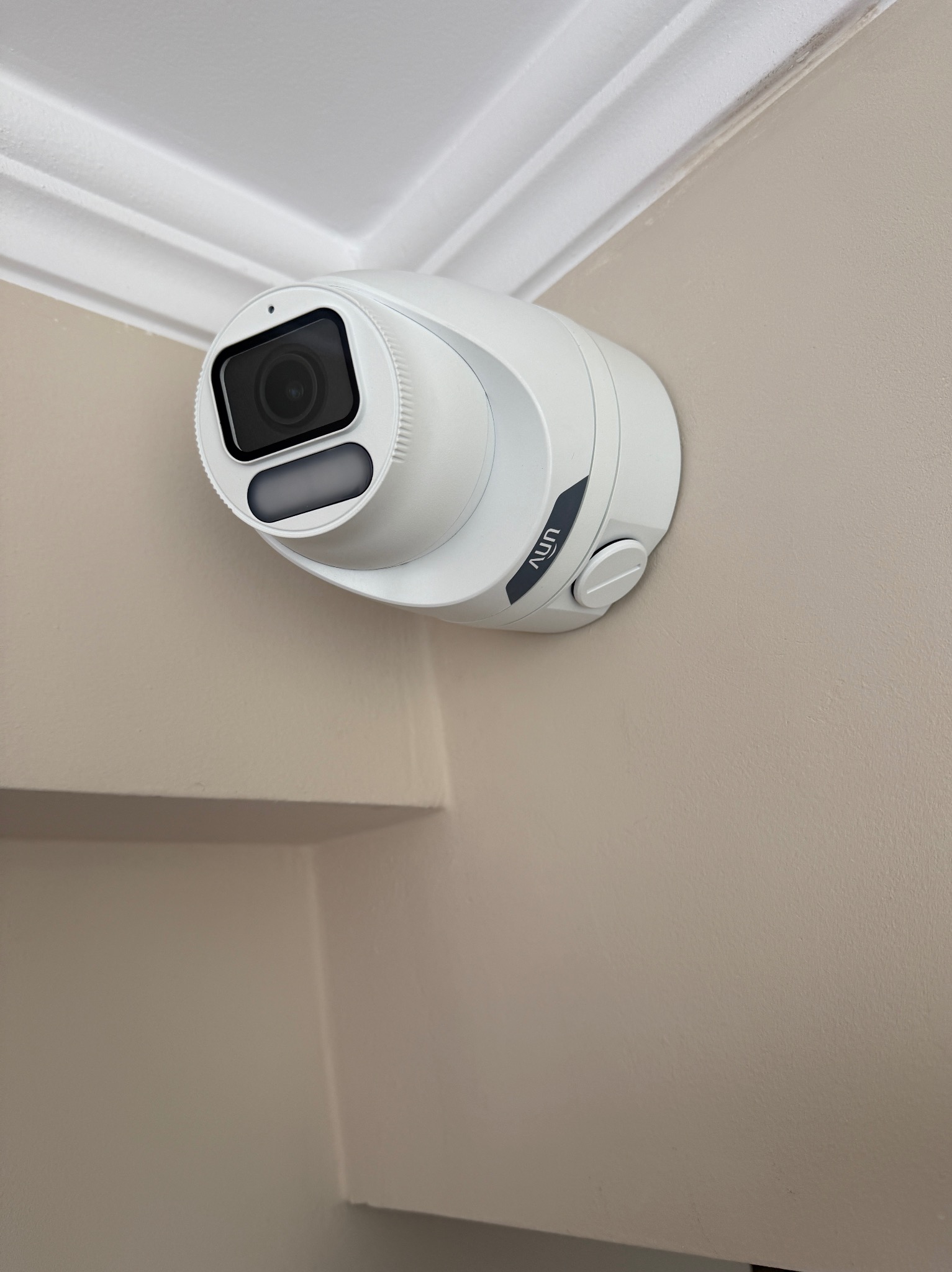 Kamera monitoringu UNV zamontowana w rogu pomieszczenia, widoczna na tle jasnej ściany i białego sufitu z ozdobnym gzymsem.