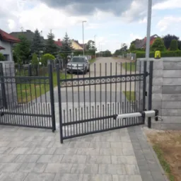 Otwarta, metalowa brama wjazdowa z automatyką, zamontowana na murowanych słupkach z szarej kostki, prowadząca do posesji z szarą kostką brukową i widocznym samochodem osobowym w tle.