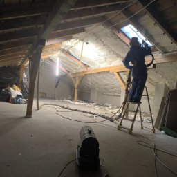 Hubert Gurzyński PUR-DOM - Pracownik w kombinezonie na drabinie aplikuje izolację natryskową na poddasze. Widoczne drewniane belki, oświetlenie i sprzęt do natrysku.